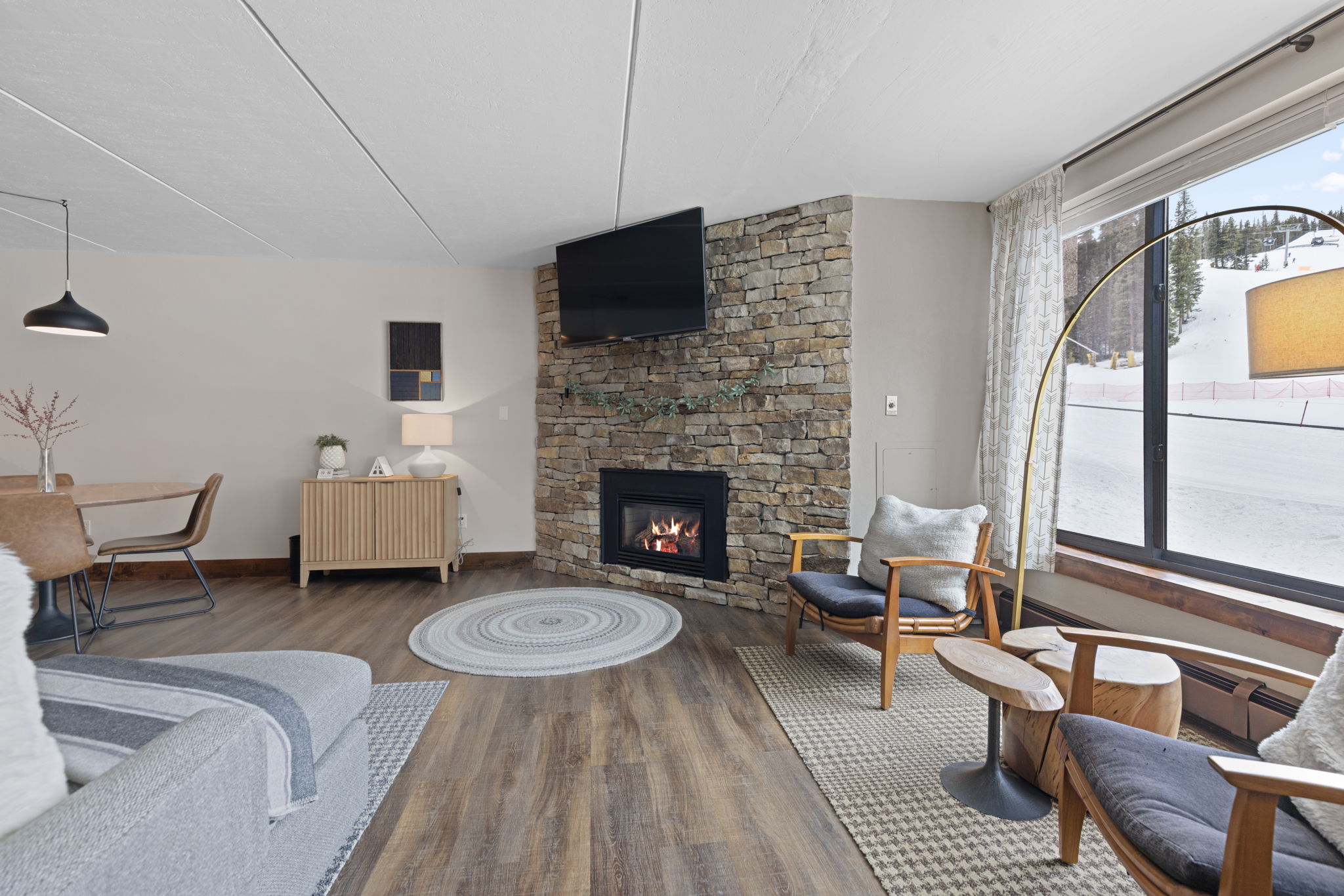 This cozy, mountain-inspired living room features a striking stone fireplace as its focal point, topped with a wall-mounted television. The space is furnished with comfortable seating, including a grey sofa and wooden armchairs, all arranged on textured area rugs over wood-look flooring. A large window provides a scenic view of a snowy ski slope, creating a warm and inviting atmosphere perfect for a winter retreat.
