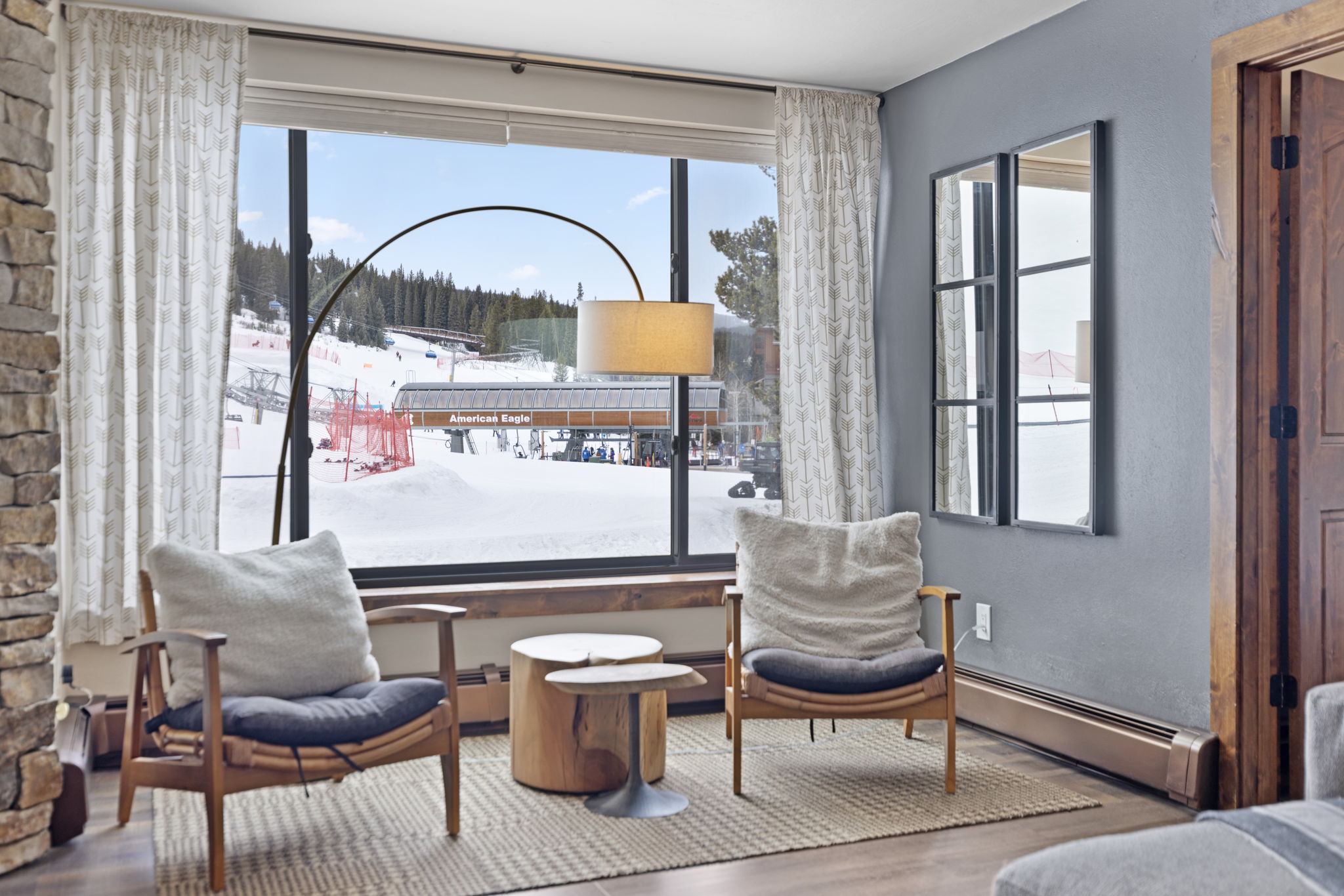 This cozy living area features two mid-century modern wooden armchairs with plush cushions, centered around a unique tiered wooden stump coffee table. A large, floor-to-ceiling window serves as the focal point, offering a stunning, direct view of a snowy ski resort and the American Eagle ski lift. The room is accented by a tall, elegant arc floor lamp and a decorative wall mirror, creating a warm, inviting atmosphere perfect for après-ski relaxation.