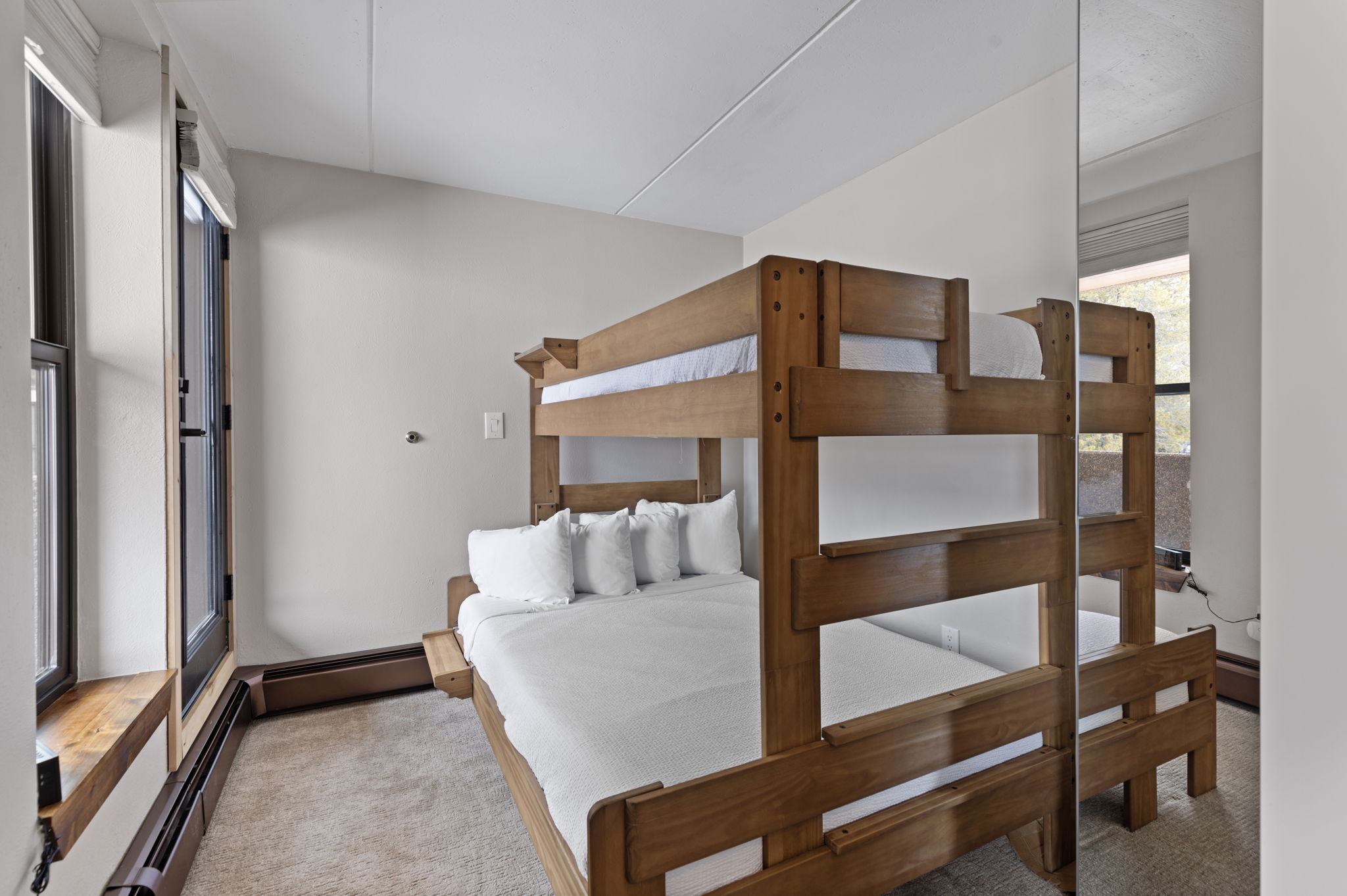 This bedroom features a sturdy wooden bunk bed with a full-sized mattress on the bottom and a twin on top, dressed in crisp white linens. The room is accented by a large floor-to-ceiling mirror that reflects the bed and a window, creating a sense of added depth and brightness. A window with a wooden sill sits to the left, providing natural light and a view of the outdoors, while the neutral wall color keeps the space feeling clean and inviting.