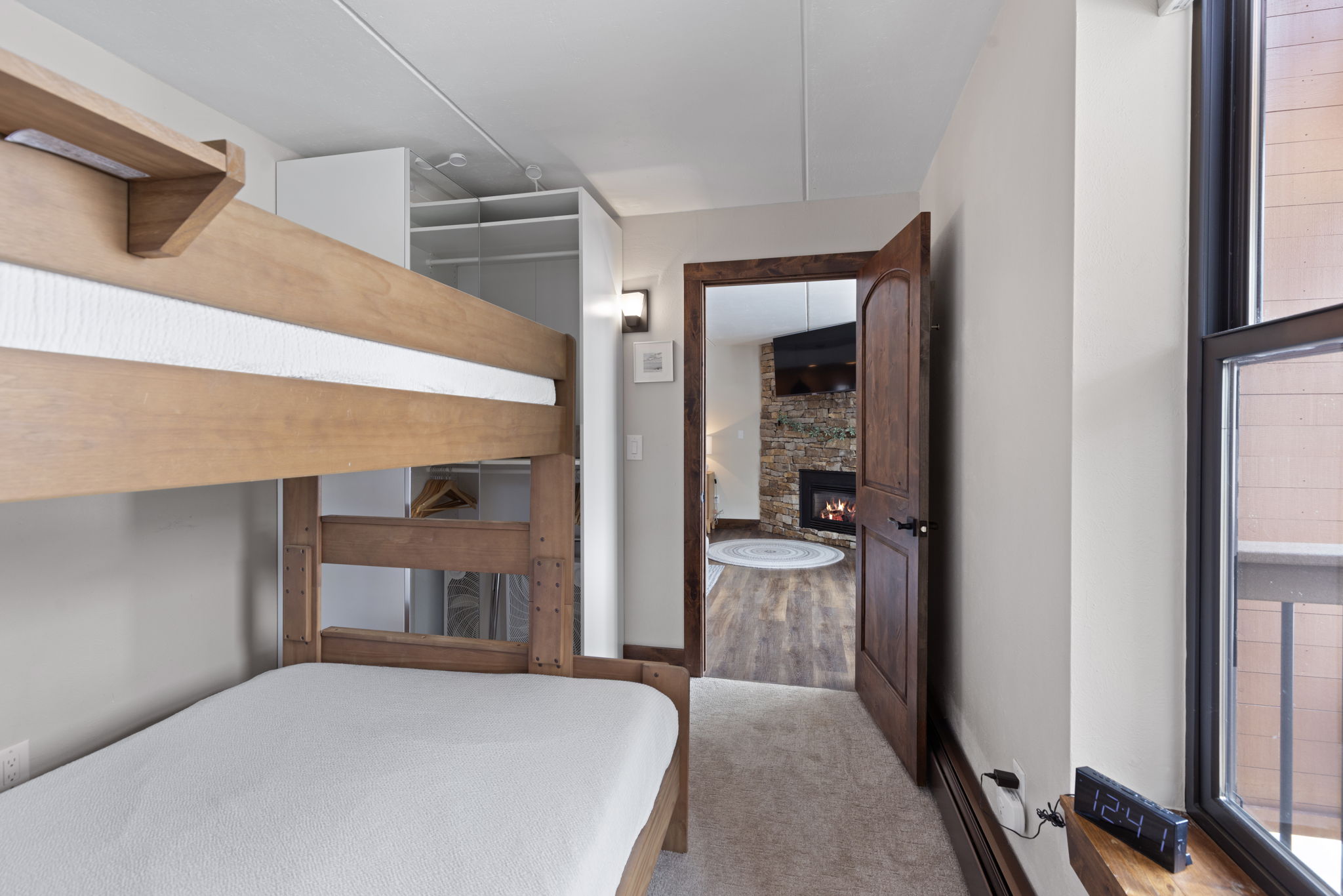 This cozy bedroom features a wooden bunk bed and a built-in white wardrobe, creating a functional space for guests or children. A doorway leads into an adjacent living area, showcasing a stone fireplace and a television, which adds a warm and inviting atmosphere to the suite. The perspective from the bedroom looking out provides a seamless transition between the sleeping quarters and the main living space.