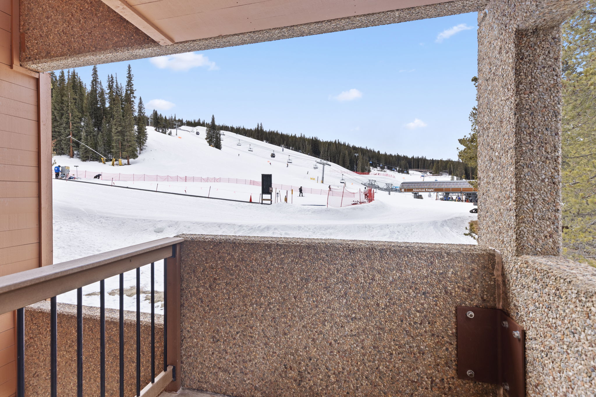This image captures a balcony view overlooking a snowy ski slope, framed by a textured stone wall and a wooden railing. The perspective provides a direct, elevated look at the ski lift and mountain terrain, emphasizing the property's prime ski-in/ski-out location. The bright, clear sky and pristine snow create an inviting and adventurous atmosphere.