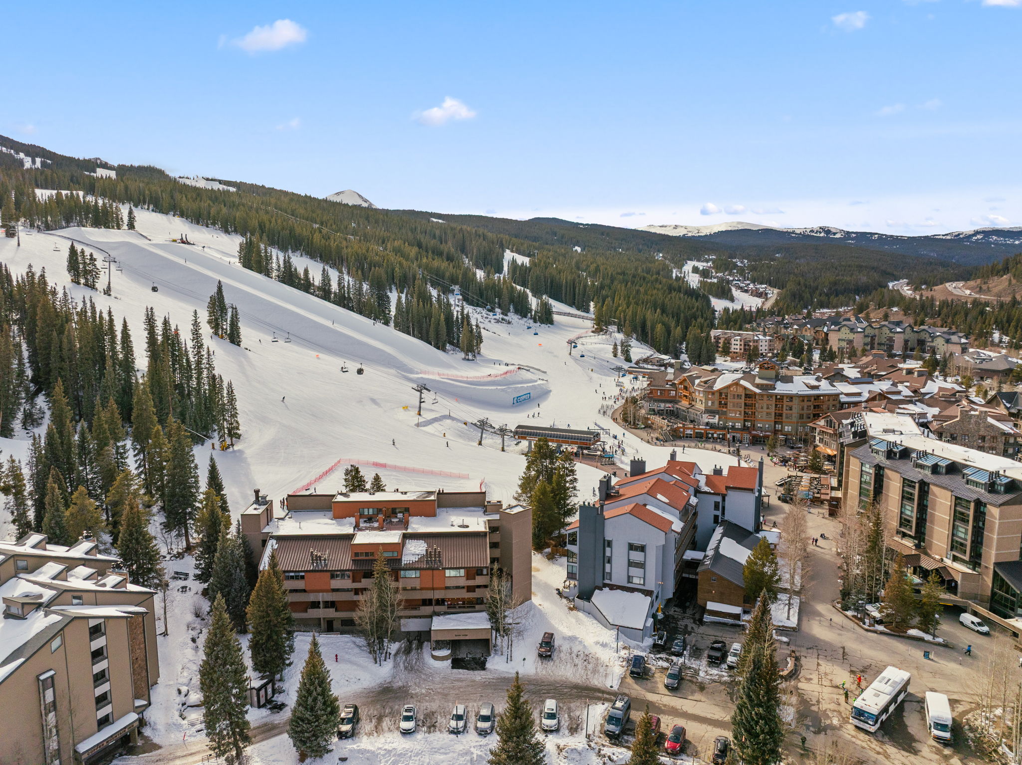 This high-angle aerial view captures a vibrant ski resort village nestled at the base of a snow-covered mountain slope. The scene features multi-story lodge-style buildings with distinct architectural designs, surrounded by evergreen trees and ski runs, creating a picturesque winter destination. The perspective emphasizes the proximity of the accommodations to the ski lifts and the bustling activity of the resort area.