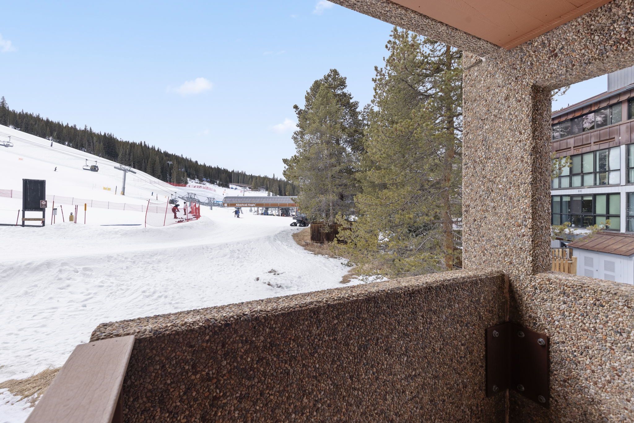 This image captures a view from a balcony featuring a textured, aggregate concrete railing that overlooks a snowy ski slope. The perspective highlights the proximity to the ski lift and mountain terrain, emphasizing the property's prime location for winter sports enthusiasts. The scene is bright and clear, showcasing the convenience of ski-in/ski-out access.