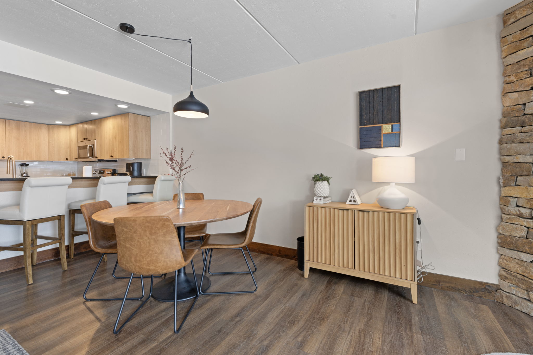 This inviting dining area features a warm, mid-century modern aesthetic with a round wooden table surrounded by four tan leather chairs. To the right, a fluted wooden sideboard topped with a lamp and decorative accents sits against a neutral wall, while a stone-clad pillar adds a rustic texture to the space. The open-concept design flows seamlessly into a bright kitchen with light wood cabinetry and a breakfast bar, creating a cohesive and functional living environment.