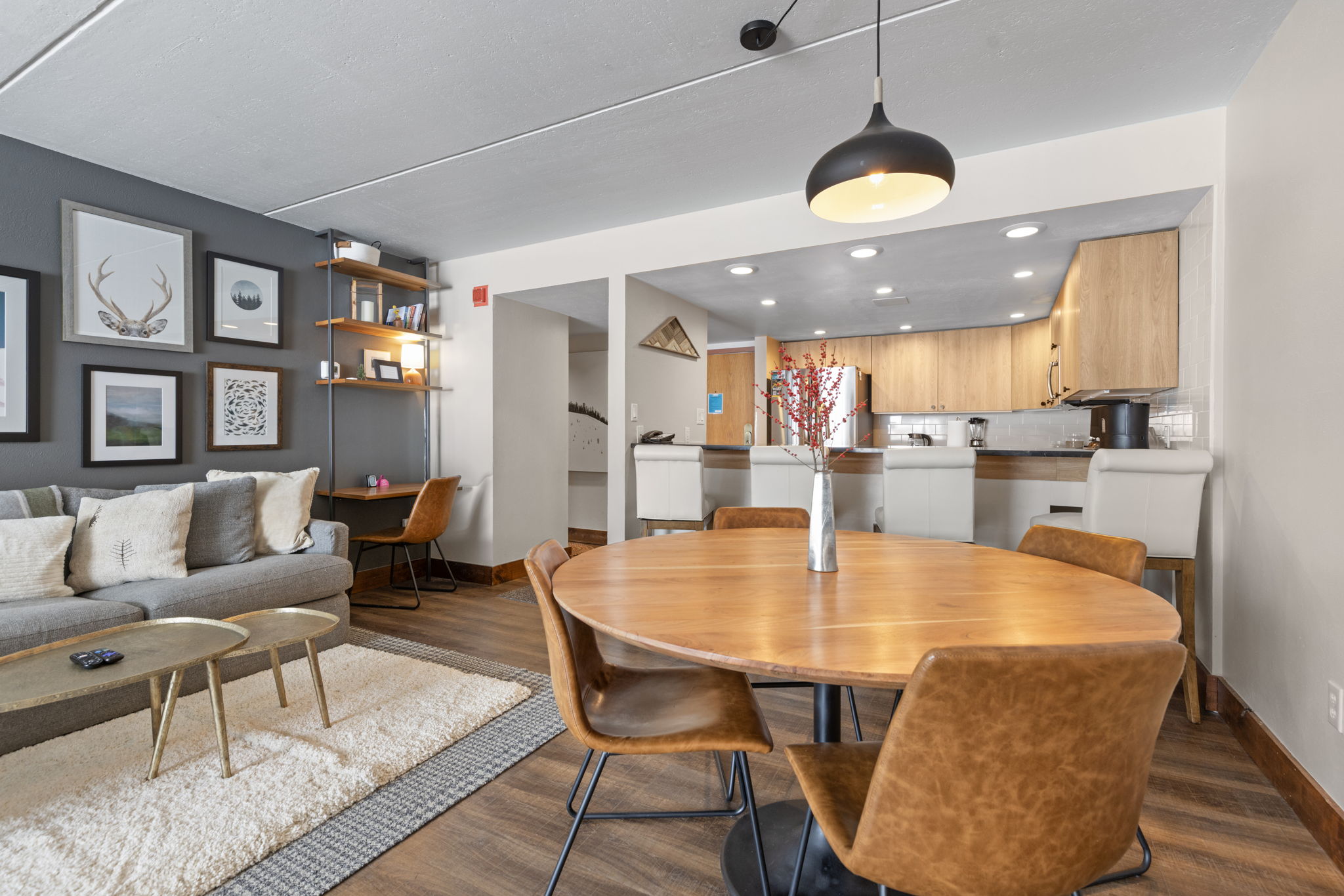 This open-concept living and dining area features a modern, warm aesthetic with wood-look flooring and a neutral color palette. The space includes a cozy gray sofa, a round wooden dining table with leather chairs, and a dedicated workspace with open shelving, all illuminated by a stylish pendant light. The perspective captures the seamless flow between the living area and the kitchen, highlighting a functional and inviting home layout.