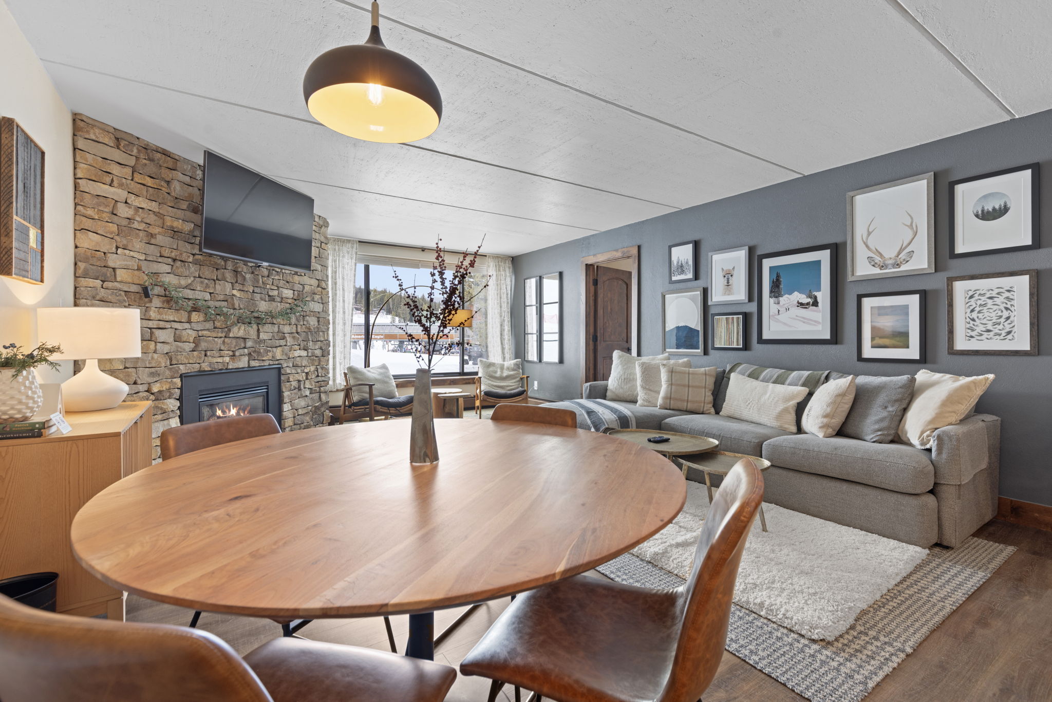 This open-concept living and dining area features a warm, rustic aesthetic with a prominent stone fireplace, a large round wooden dining table, and a comfortable grey sectional sofa. The space is accented by a gallery wall of framed art, a modern pendant light, and large windows that provide a scenic view of a snowy landscape. The combination of natural textures and cozy furnishings creates an inviting, mountain-lodge atmosphere perfect for relaxation.