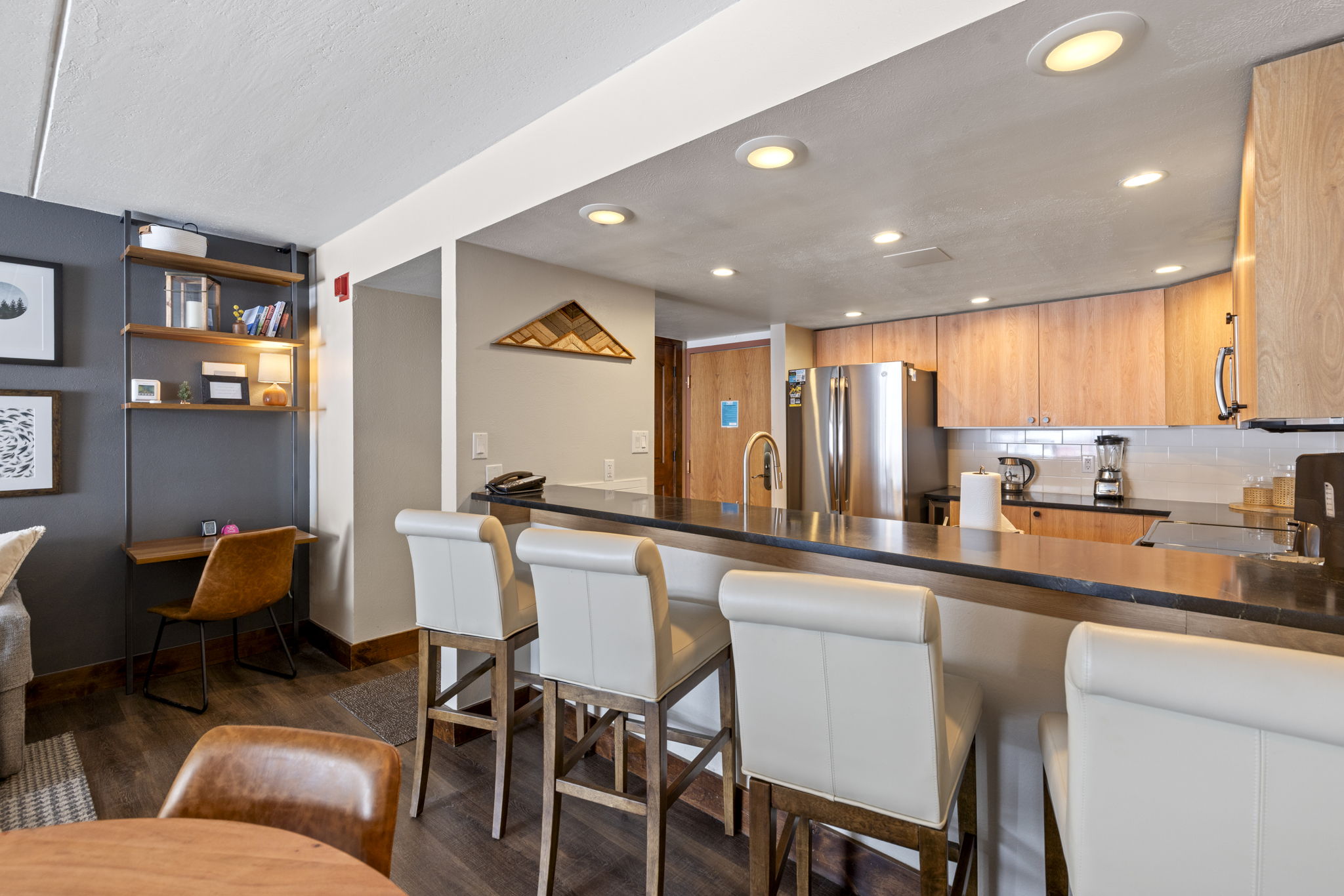 This open-concept kitchen features a sleek breakfast bar with four modern cream-colored bar stools, perfect for casual dining. The space is highlighted by warm wood cabinetry, stainless steel appliances, and recessed lighting, creating a welcoming and functional atmosphere. A dedicated workspace with open shelving is integrated into the living area, showcasing a thoughtful and efficient use of space.