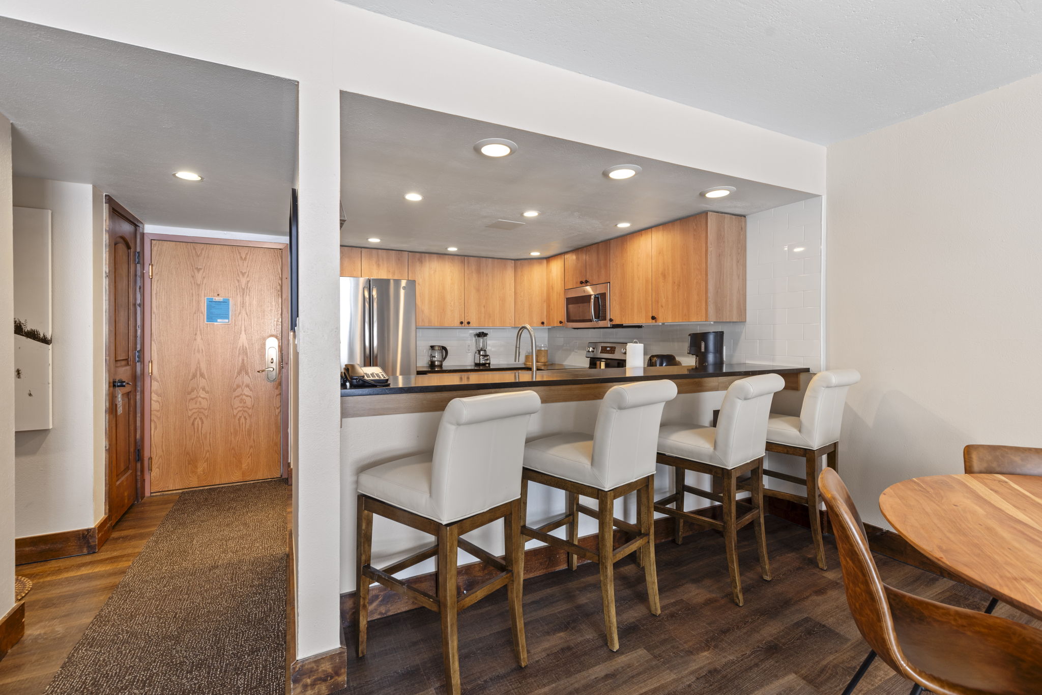 This open-concept kitchen features warm wood cabinetry, stainless steel appliances, and a breakfast bar with four upholstered stools. The space flows seamlessly into an entryway and dining area, creating a functional and inviting layout. The warm wood tones and neutral color palette provide a cozy, well-maintained atmosphere suitable for a vacation rental or home.