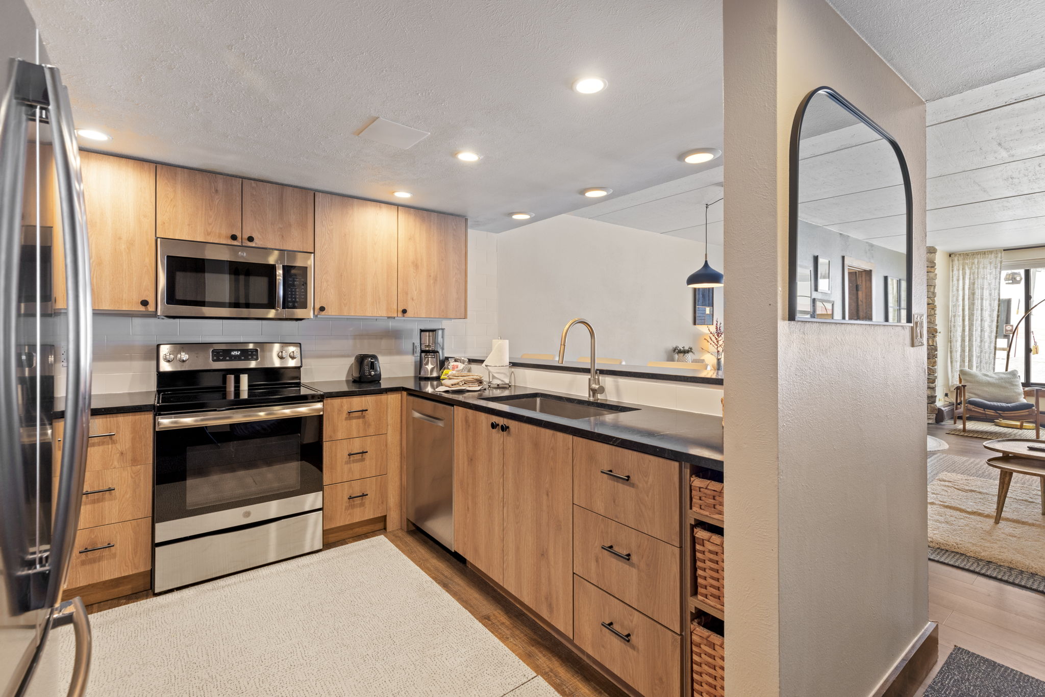 This modern kitchen features warm wood-toned cabinetry paired with sleek black countertops and stainless steel appliances, creating a contemporary and inviting aesthetic. The open-concept layout includes a pass-through window that connects to the living area, enhancing the flow of the home. A light-colored area rug anchors the space, while recessed lighting provides a bright and functional atmosphere.