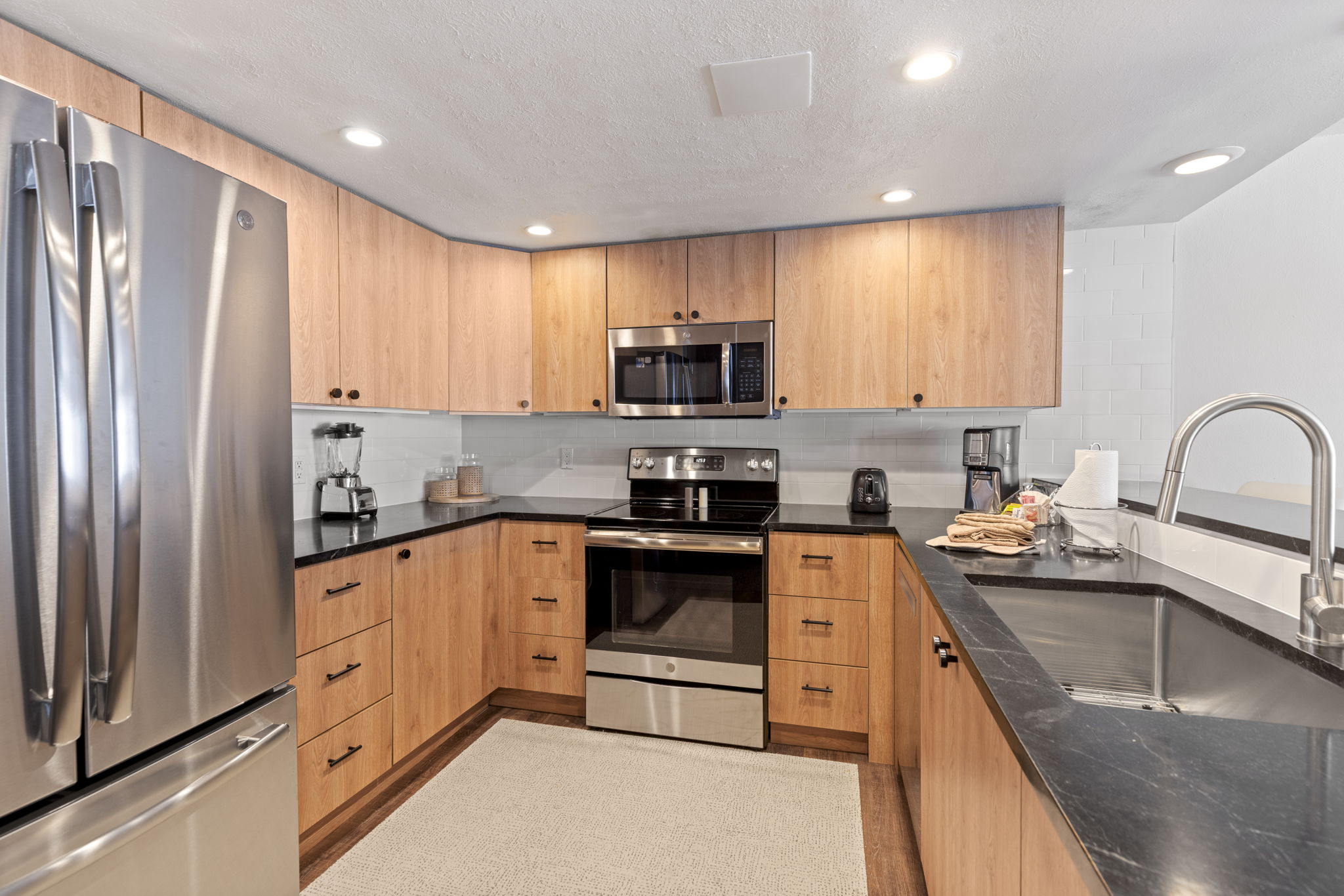 This modern kitchen features warm wood-toned cabinetry paired with sleek black countertops and a white subway tile backsplash. The space is equipped with stainless steel appliances, including a large refrigerator, a built-in microwave, and an oven range. A neutral-toned area rug sits on the floor, and the perspective is a wide-angle shot capturing the functional layout and clean, contemporary aesthetic.