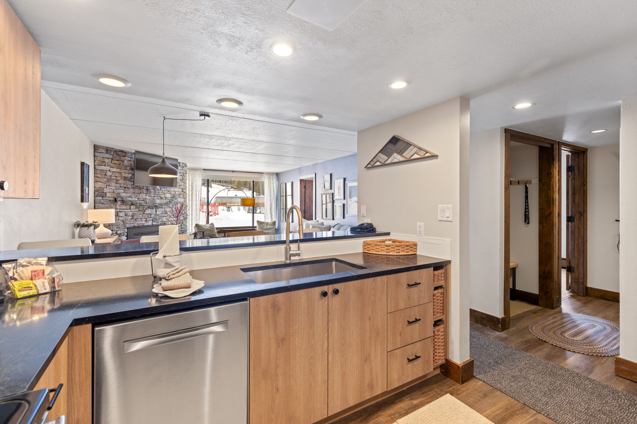 This modern kitchen features warm wood-toned cabinetry paired with sleek black countertops and stainless steel appliances. The open-concept design allows for a clear view into the living area, which is anchored by a rustic stone fireplace and large windows overlooking a snowy landscape. The space feels cozy and inviting, blending contemporary finishes with a comfortable, mountain-inspired aesthetic.