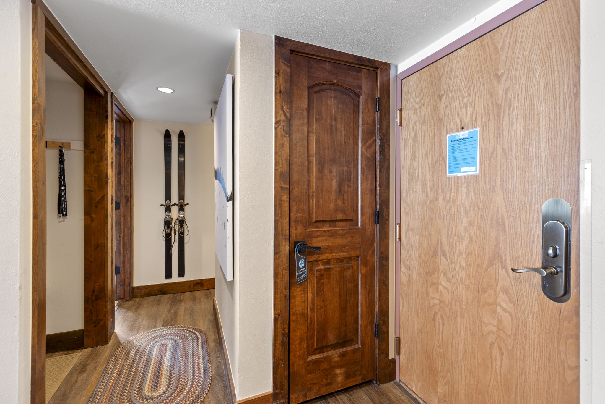 This interior hallway features warm wood-toned flooring and matching dark wood door frames, creating a cozy, rustic mountain aesthetic. The space includes a wooden door with a modern electronic lock, a decorative wall-mounted ski display, and a braided oval rug that adds texture to the entryway. The perspective is a straight-on view looking down the corridor, highlighting the transition between the entrance and the rest of the home.