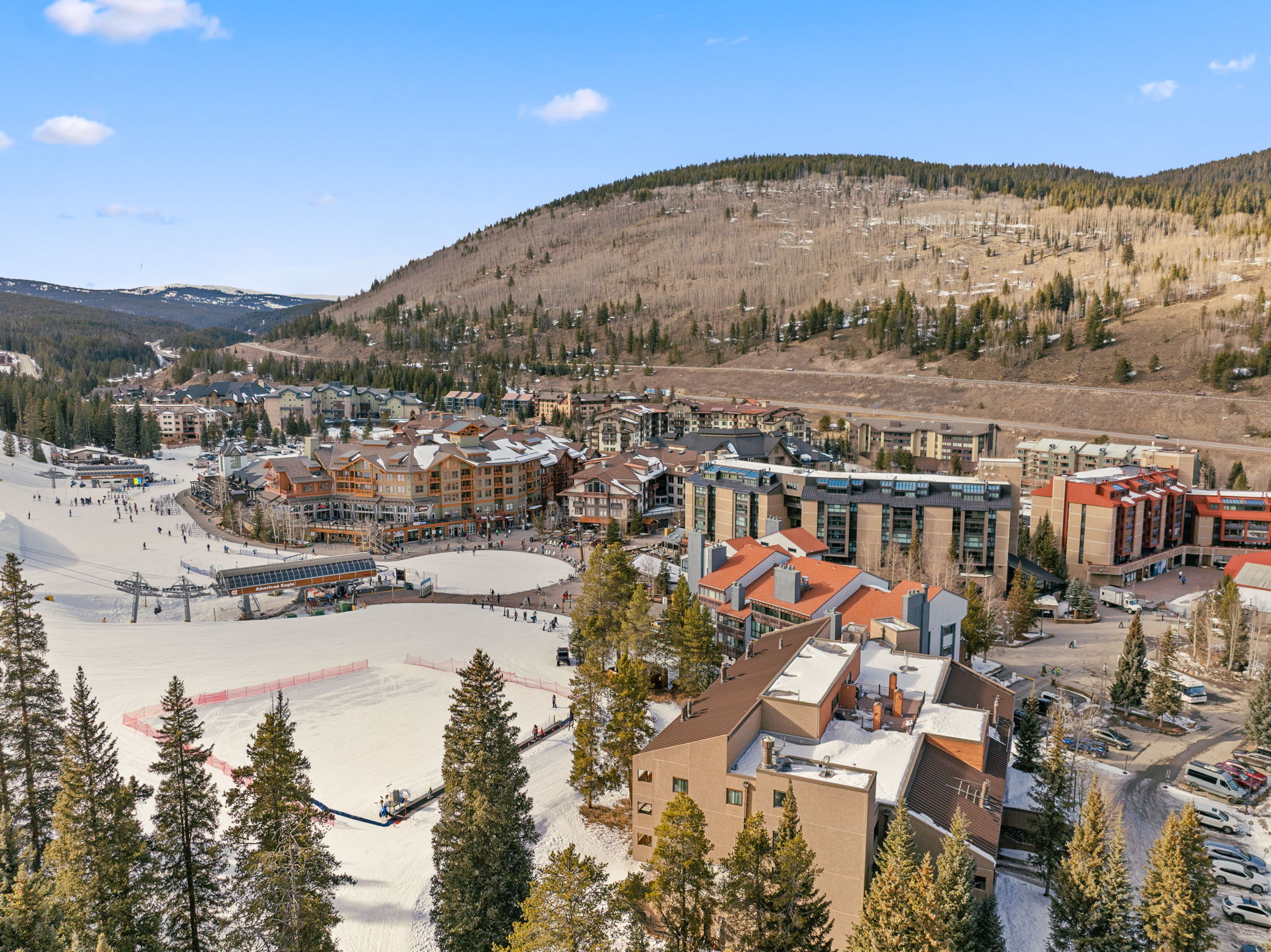 This high-angle aerial view captures a vibrant ski resort village nestled at the base of a snow-dusted mountain range. The scene features a mix of multi-story lodges with classic alpine architecture, a prominent ski lift, and a central plaza area surrounded by evergreen trees. The bright, clear sky and snowy terrain create an inviting atmosphere, highlighting the resort's prime location for winter sports and mountain recreation.