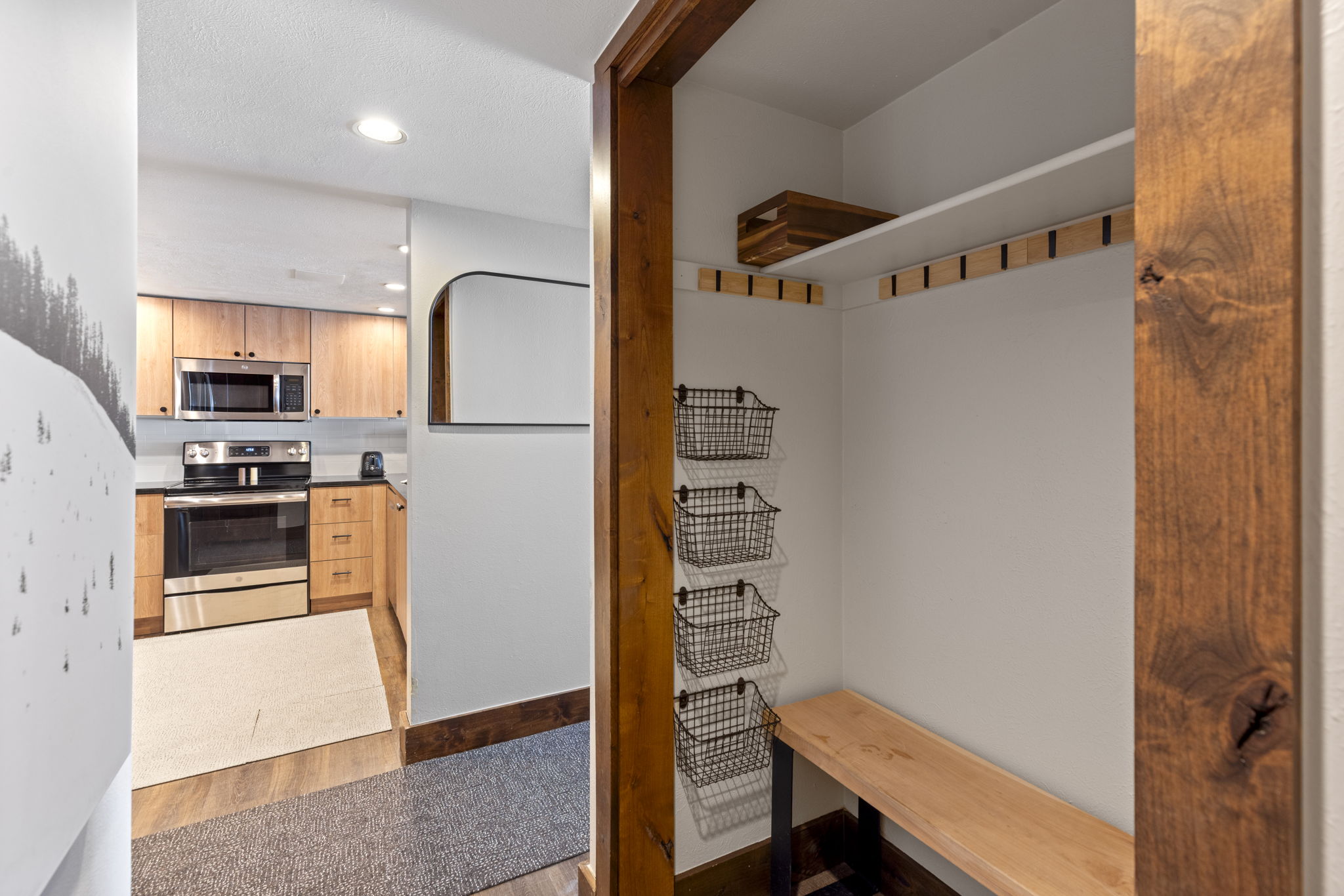 This perspective captures a transition space featuring a mudroom area with a wooden bench, wall-mounted wire storage baskets, and coat hooks, leading into a bright, modern kitchen. The kitchen is equipped with light wood cabinetry, stainless steel appliances, and a clean, functional layout. The contrast between the warm wood tones of the mudroom and the sleek kitchen creates a welcoming and organized atmosphere.