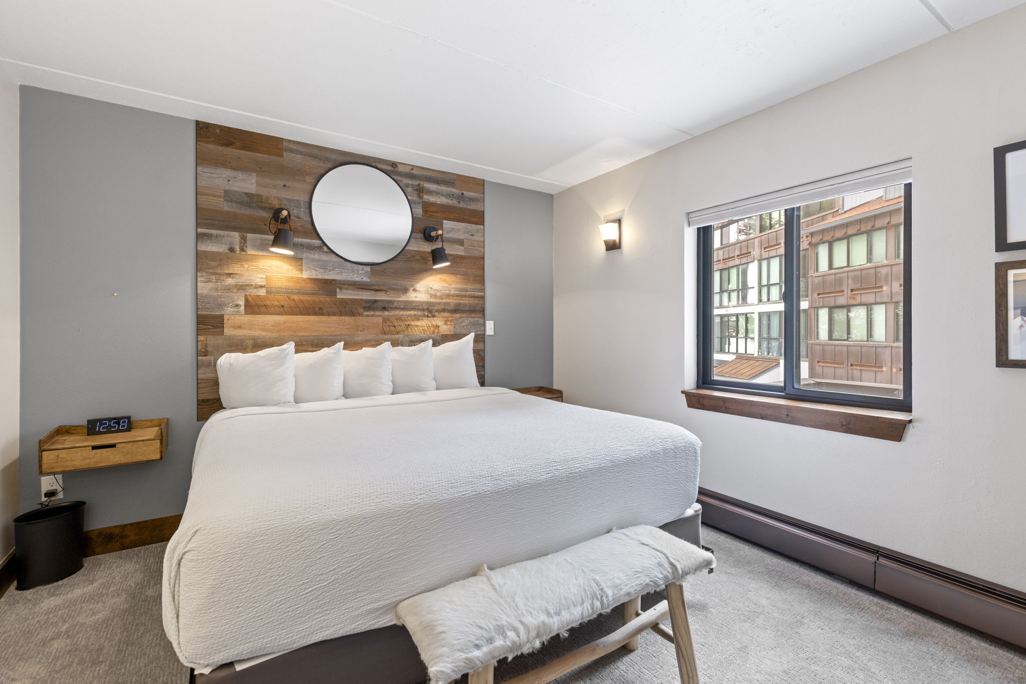 This cozy primary bedroom features a king-sized bed centered against a rustic wood-paneled accent wall with a circular mirror and dual wall-mounted sconces. A floating wooden nightstand with a digital clock adds a modern touch, while a plush white bench sits at the foot of the bed. The room is brightened by a large window that offers a view of the surrounding architecture, creating a warm and inviting atmosphere.