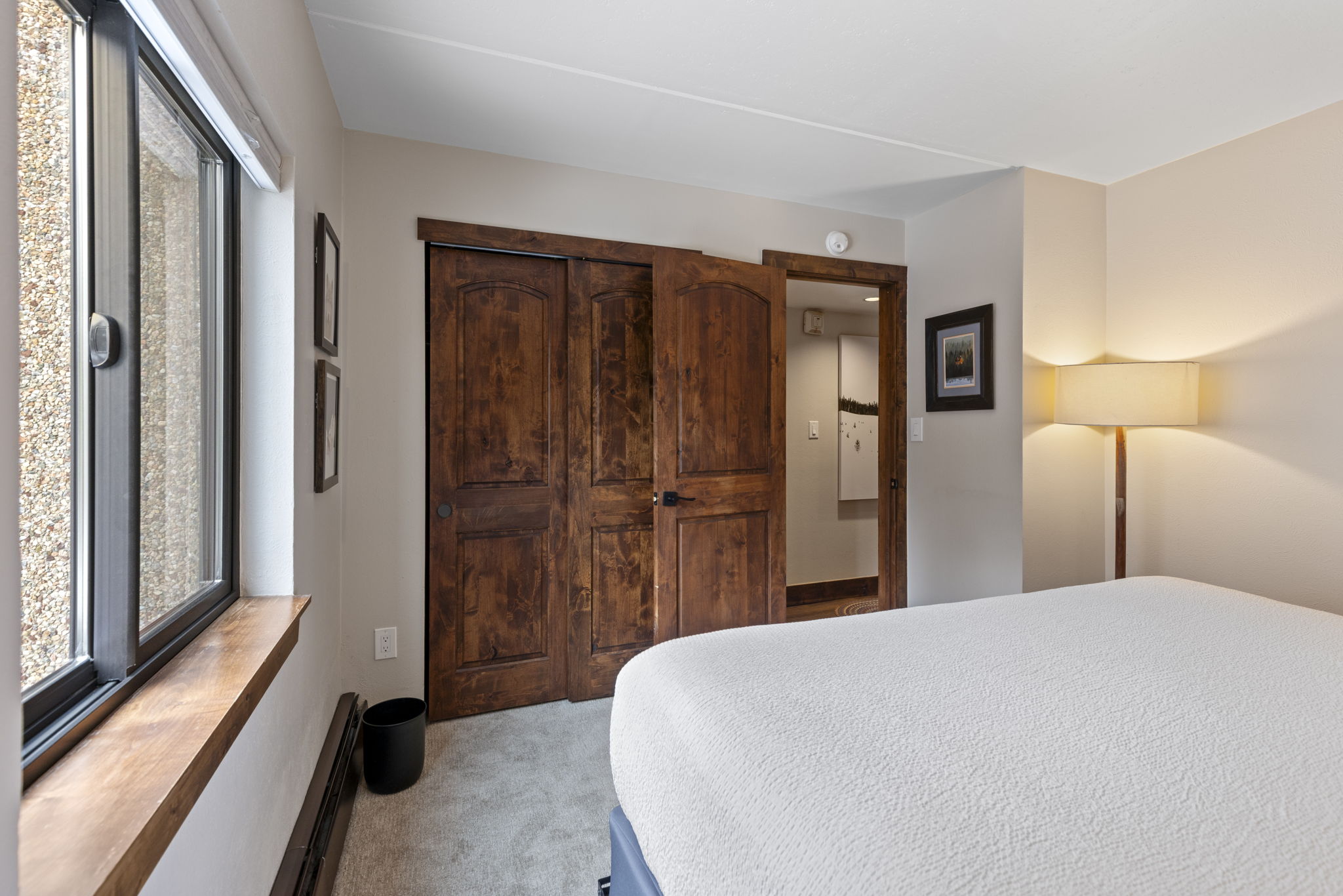 This cozy bedroom features a neutral color palette, a plush white bed, and a large window with a wooden sill that brings in natural light. The room is accented by rustic wooden closet doors and a warm floor lamp, creating a comfortable and inviting atmosphere. The perspective is from the foot of the bed, looking toward the closet and the adjacent hallway.