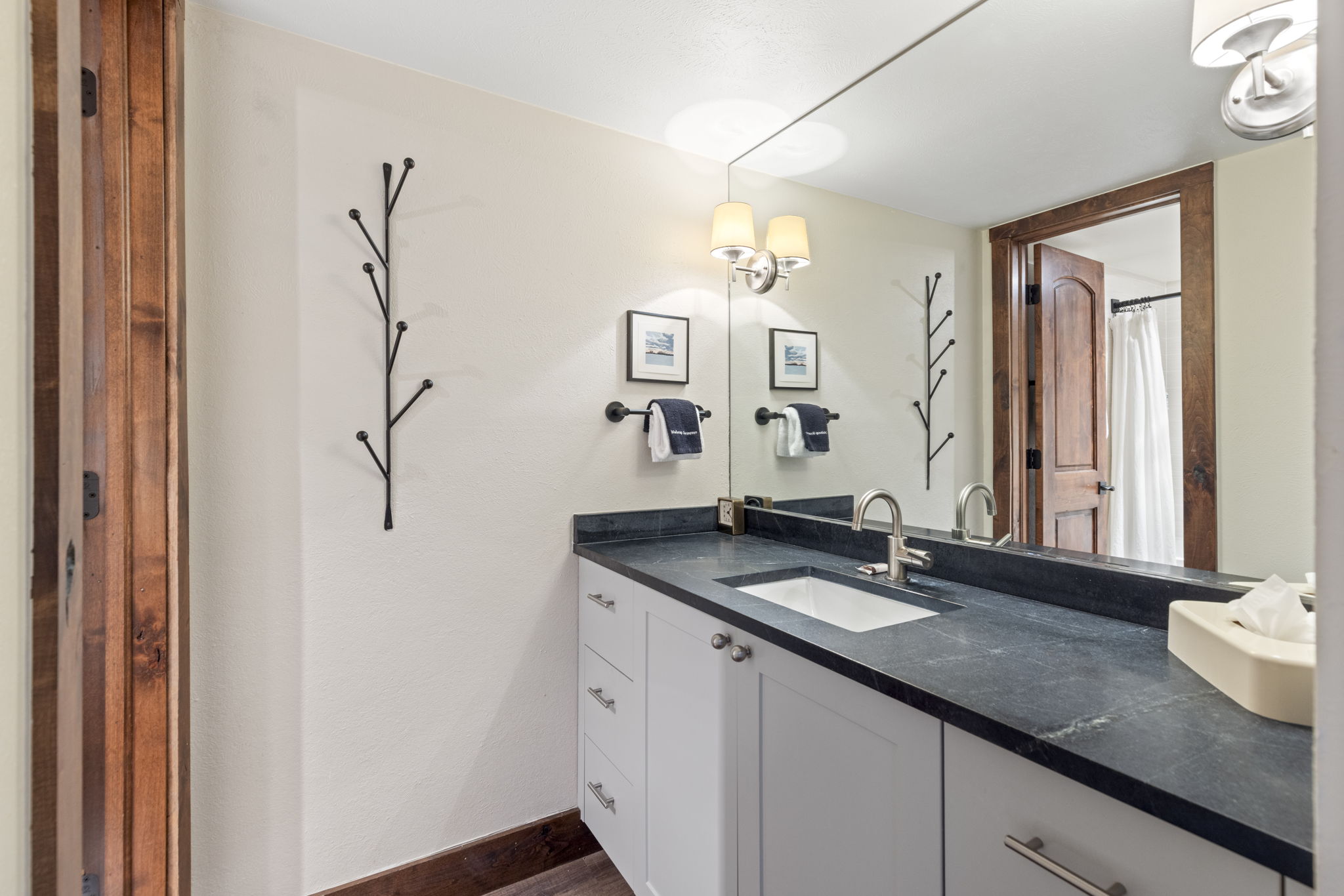 This bathroom features a sleek, modern vanity with a dark stone countertop and a white undermount sink, contrasted by light gray cabinetry. A large wall-to-wall mirror reflects the space, while a unique branch-style wall hook adds a rustic, artistic touch to the neutral-toned walls. The warm wood trim around the doorway and baseboards provides a cozy, cabin-like aesthetic that complements the contemporary fixtures.