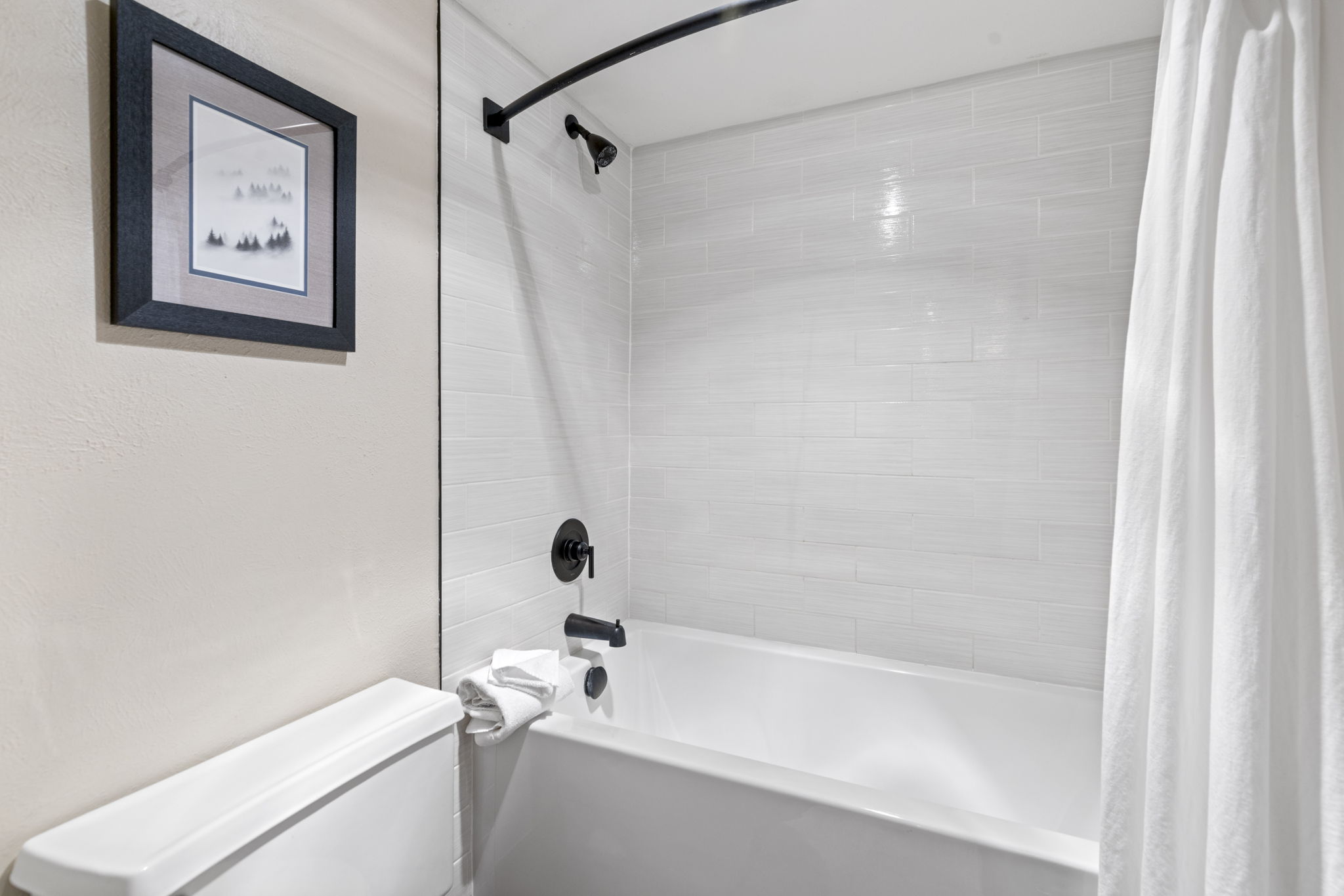 This clean and modern bathroom features a white bathtub with a sleek black shower head and faucet set against a backdrop of light-colored, horizontal subway tiles. A white shower curtain hangs from a curved black rod, and a framed piece of art adds a touch of personality to the adjacent wall. The space is bright and functional, showcasing a contemporary aesthetic suitable for a guest or secondary bathroom.