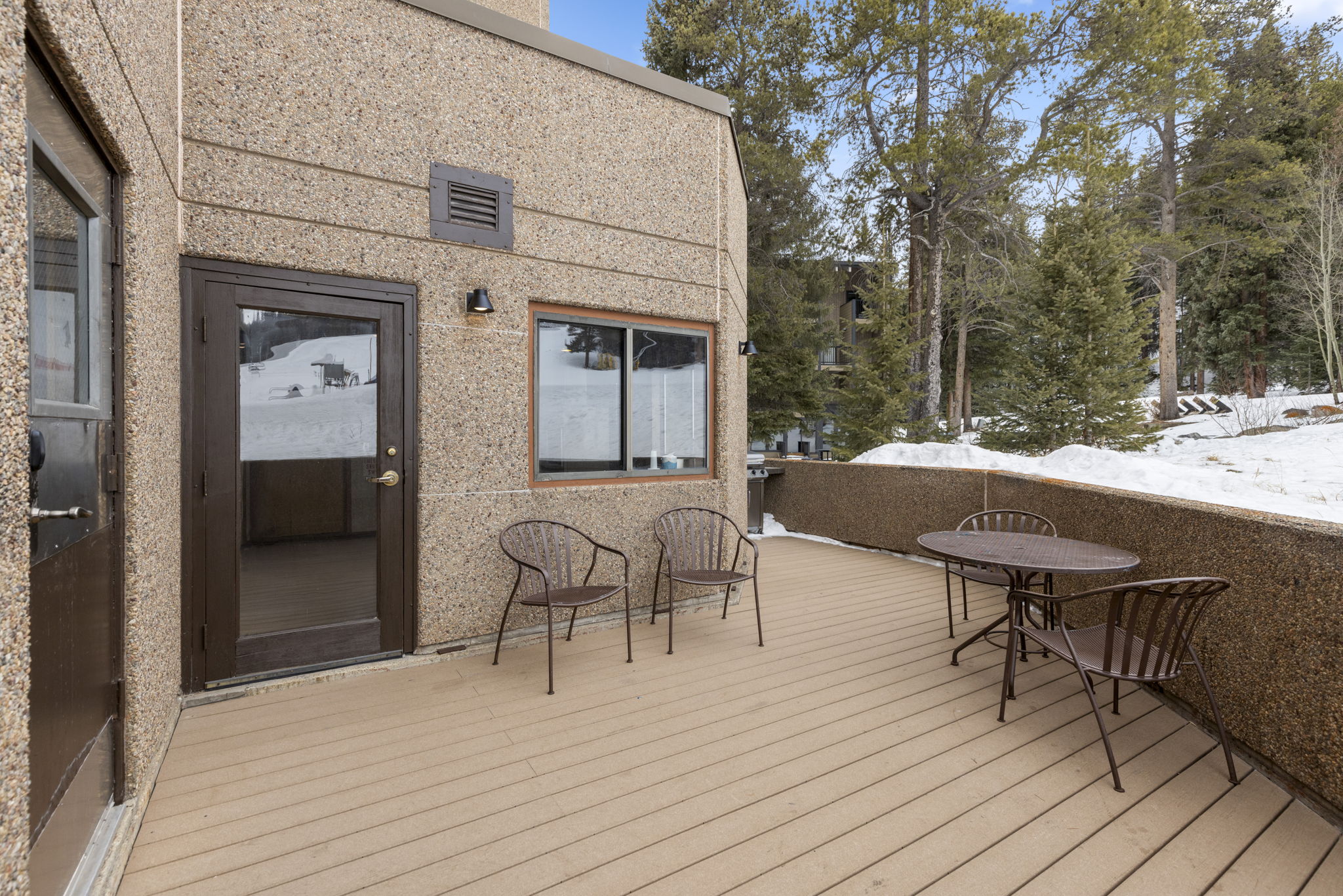 This image showcases a spacious, elevated deck featuring a textured, light-brown composite surface and a matching low-profile privacy wall. The area is furnished with a small bistro set and two additional chairs, providing a cozy outdoor seating space overlooking a snowy, wooded landscape. The perspective is a wide-angle shot from the deck entrance, emphasizing the seamless transition between the building's textured exterior and the serene, wintry environment.