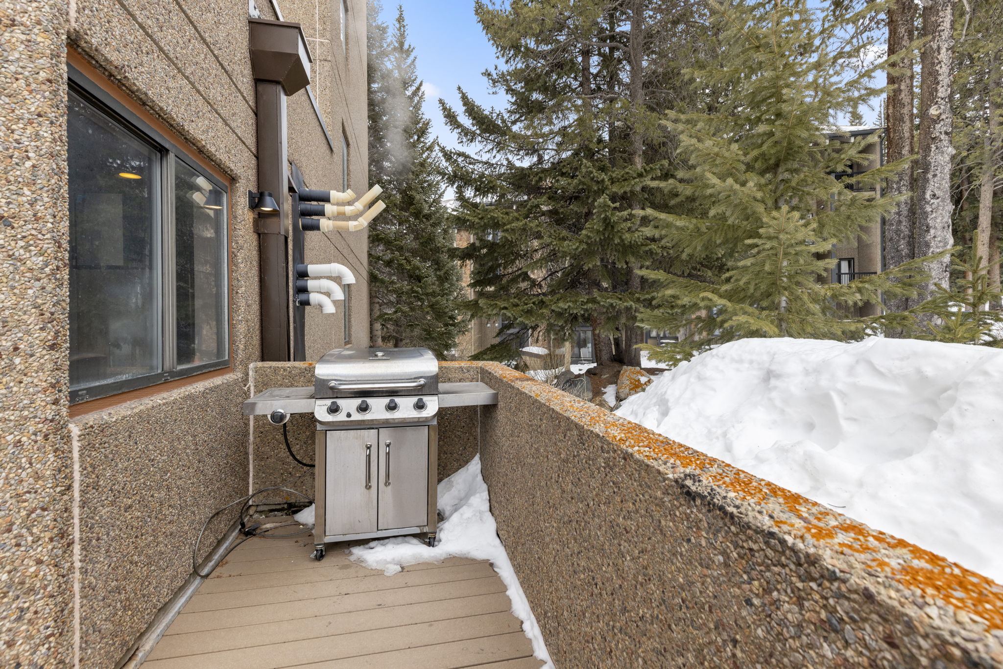 This balcony features a stainless steel gas grill set against a textured, aggregate-finished exterior wall. The deck is constructed with composite wood-look planks and overlooks a snowy, wooded landscape, creating a cozy, alpine-inspired outdoor space. The perspective captures the functional utility of the balcony while highlighting the surrounding natural environment.