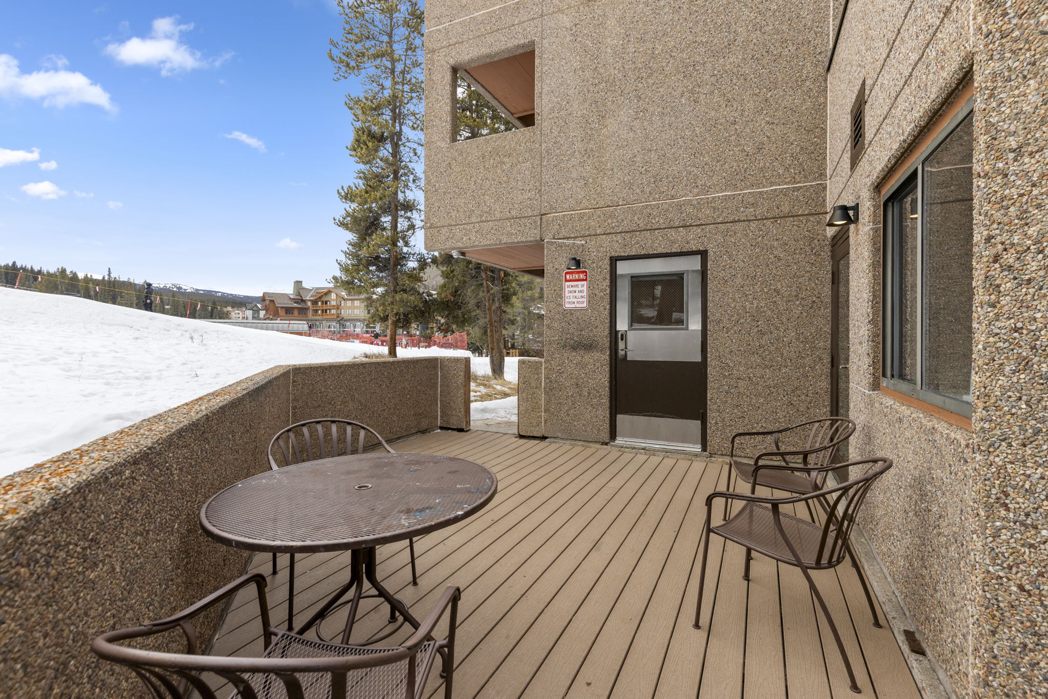 This image showcases a private, elevated deck featuring a metal patio set with a round table and three chairs, perfect for outdoor relaxation. The deck offers a direct, scenic view of a snow-covered ski slope and surrounding mountain resort buildings, emphasizing its prime location for winter sports enthusiasts. The structure is built with durable, textured concrete walls and composite decking, creating a sturdy and low-maintenance outdoor living space.