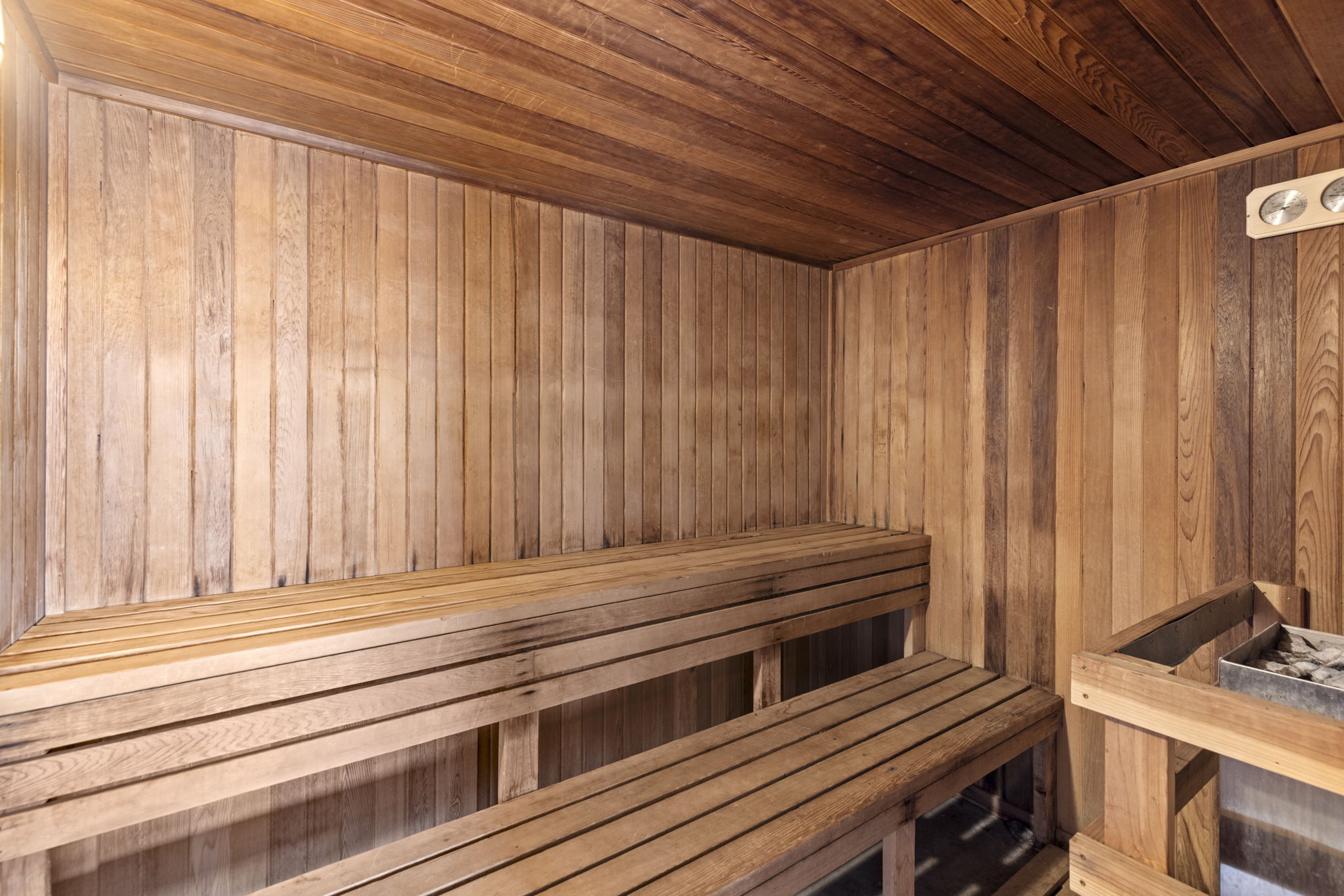 This interior view showcases a traditional wooden sauna, characterized by its warm, natural cedar paneling on the walls and ceiling. The space features tiered bench seating and a dedicated heater unit filled with stones, creating an inviting and relaxing atmosphere. The perspective is eye-level, emphasizing the craftsmanship and cozy, enclosed nature of this wellness amenity.