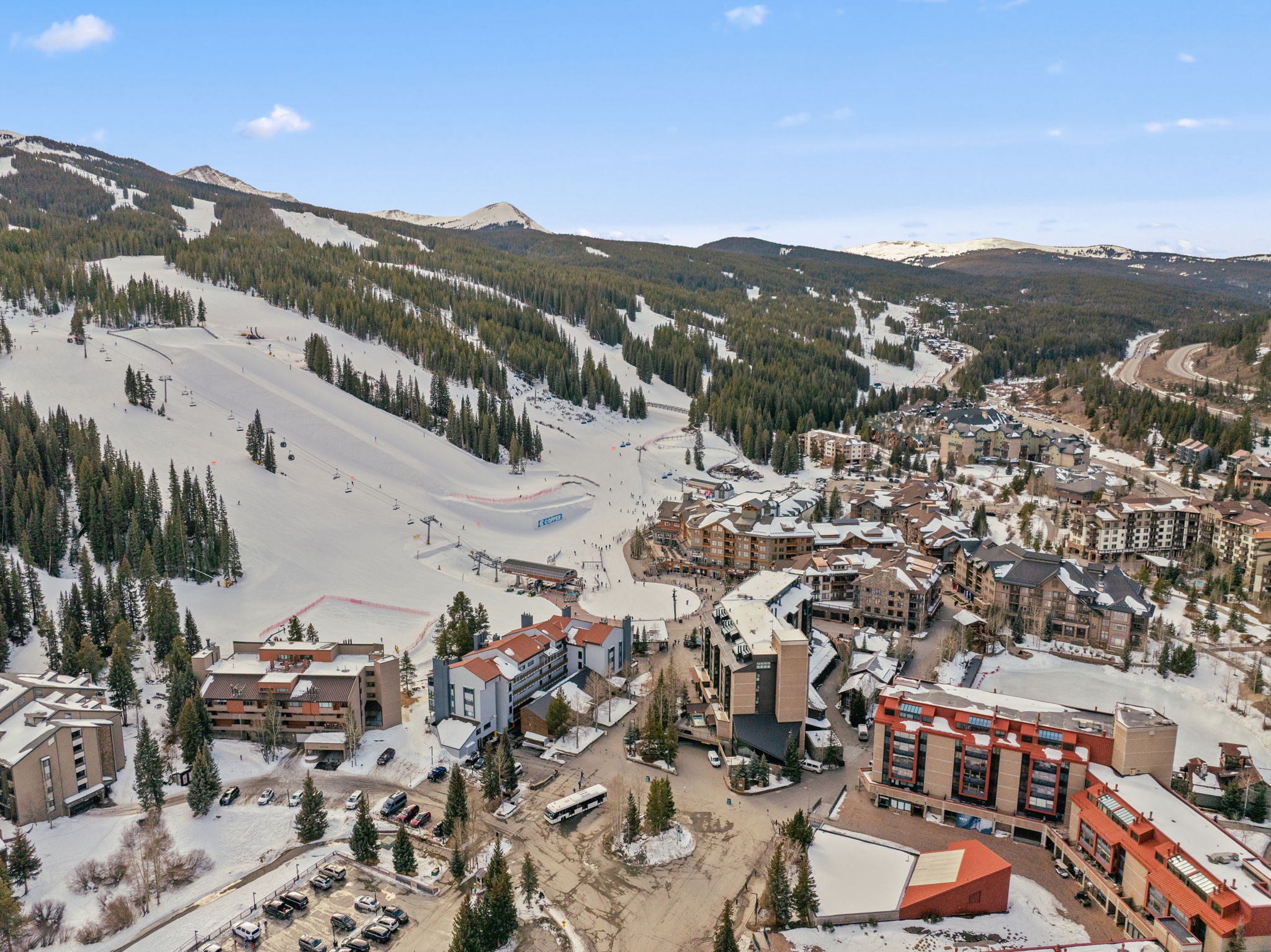 This high-angle aerial view captures a bustling ski resort village nestled at the base of a snow-covered mountain slope. The scene features multi-story lodge-style buildings with brown and grey facades, surrounded by evergreen trees and parking areas, with a clear view of the ski lifts and groomed runs ascending the mountain. The bright, clear sky and expansive landscape create an inviting atmosphere for winter sports enthusiasts and vacationers.