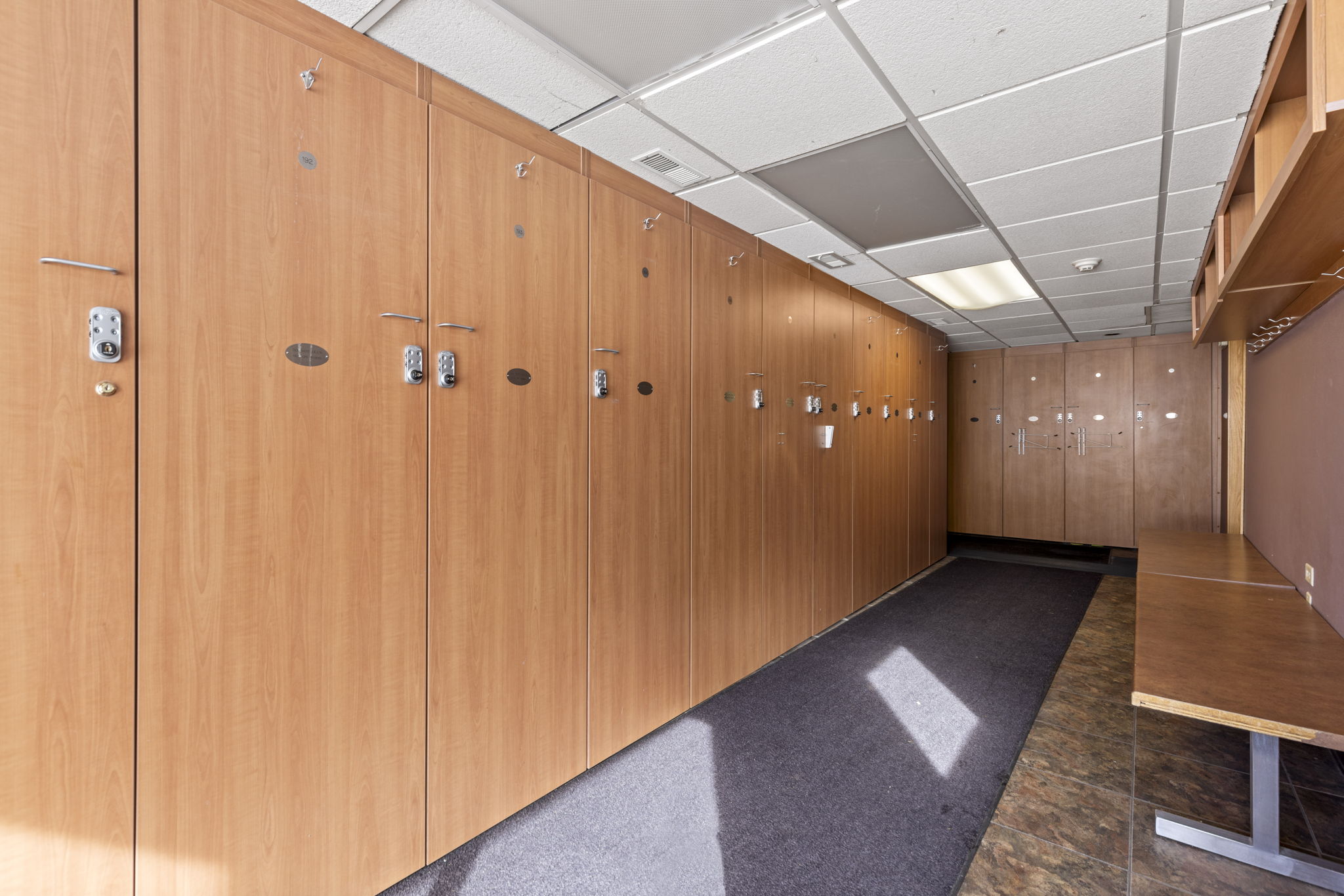 This image showcases a locker room facility, likely part of a gym or clubhouse amenity, featuring a long row of light wood-toned lockers with electronic keypads. A dark carpeted runner stretches along the floor, leading toward a wooden bench area at the end of the corridor. The space is illuminated by recessed ceiling lights and drop-ceiling panels, creating a clean, functional, and professional environment.