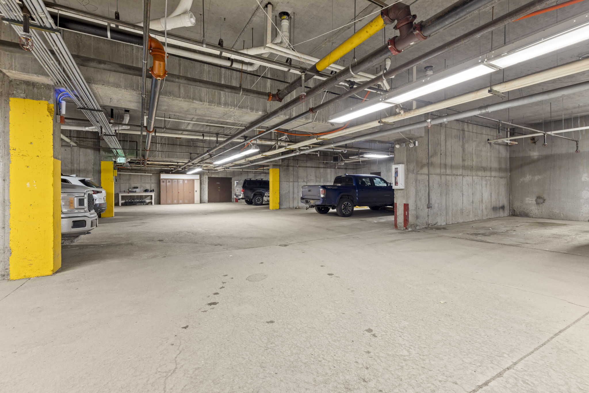 This image captures a spacious, industrial-style underground parking garage with exposed concrete ceilings and utility pipes. The area features bright yellow support pillars, fluorescent overhead lighting, and several parked vehicles, including a prominent blue pickup truck. The perspective is wide and low, emphasizing the expansive, utilitarian nature of the parking facility.