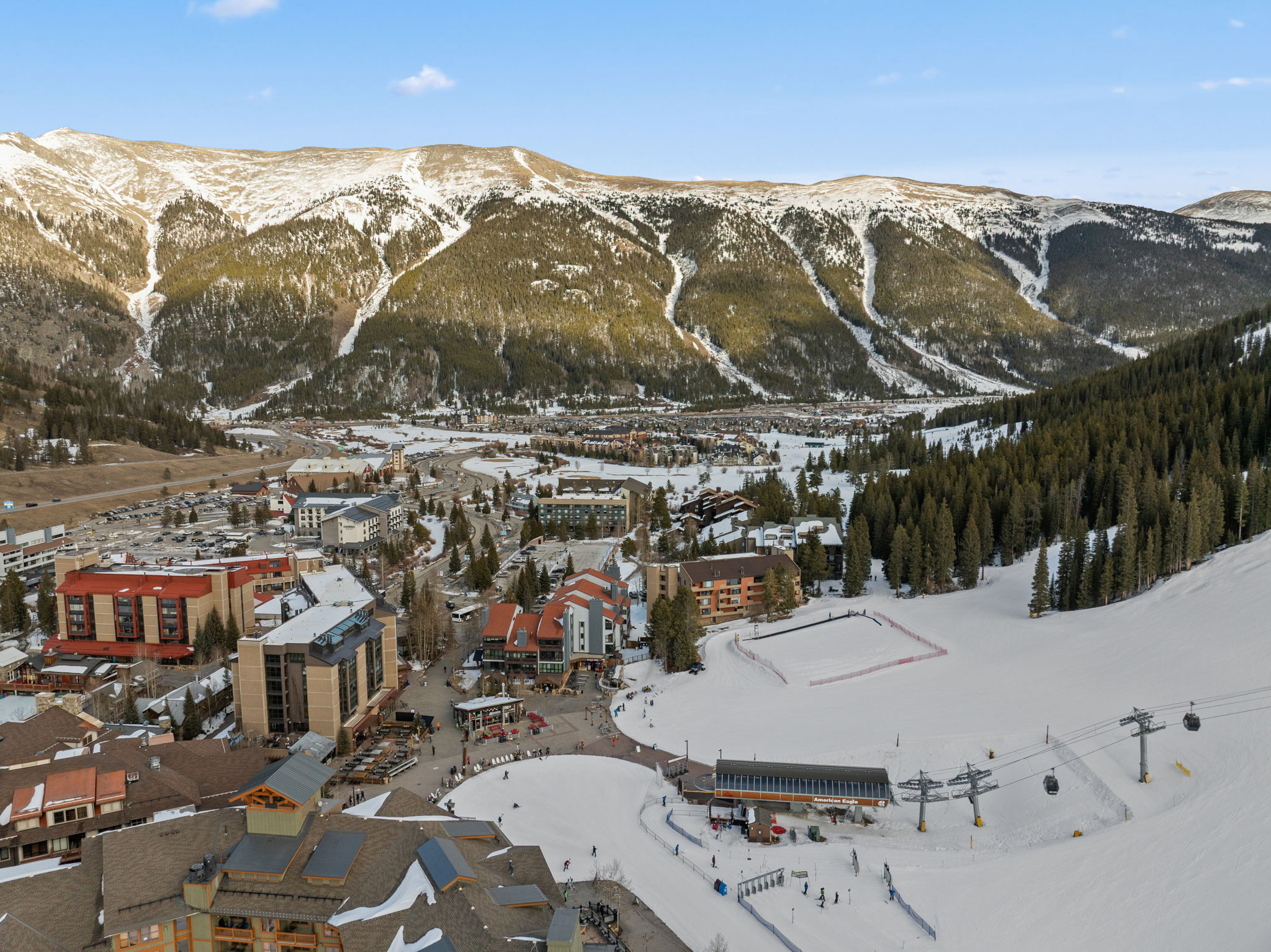 This high-angle aerial view captures a bustling ski resort village nestled at the base of a majestic, snow-capped mountain range. The scene features a mix of multi-story lodge-style buildings, a ski lift terminal, and groomed slopes, all set against a clear blue sky. The perspective provides a comprehensive overview of the resort's layout, highlighting its prime location for winter sports and mountain tourism.