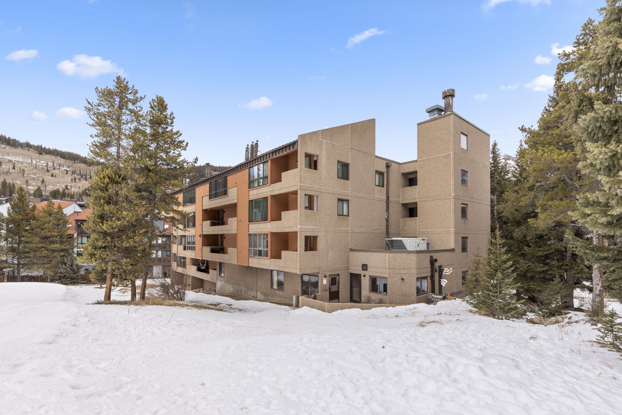 This exterior side view captures a multi-story condominium complex nestled in a snowy, mountainous landscape. The building features a mix of textured concrete and warm-toned wood siding, with stacked balconies and large windows that offer views of the surrounding pine trees and ski slopes. The perspective is from a low angle on the snow-covered ground, emphasizing the building's height and its integration into the alpine environment.