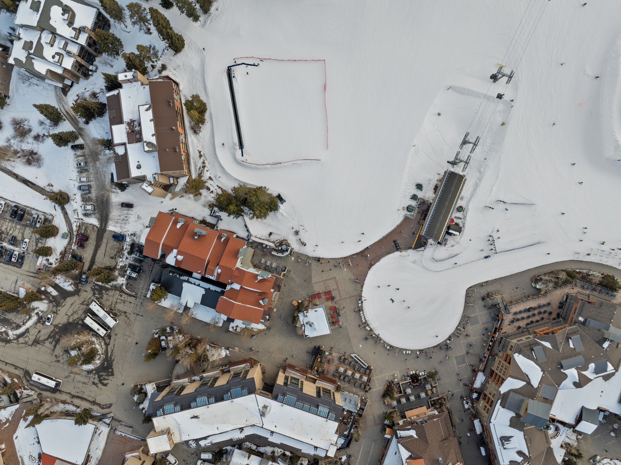 This high-angle aerial view captures a bustling ski resort village nestled in a snowy landscape. The scene features a mix of multi-story lodge-style buildings with brown roofs, a central plaza area, and a ski lift terminal adjacent to a groomed snow-covered slope. The perspective provides a comprehensive overview of the resort's layout, highlighting the integration of hospitality structures with winter recreational facilities.