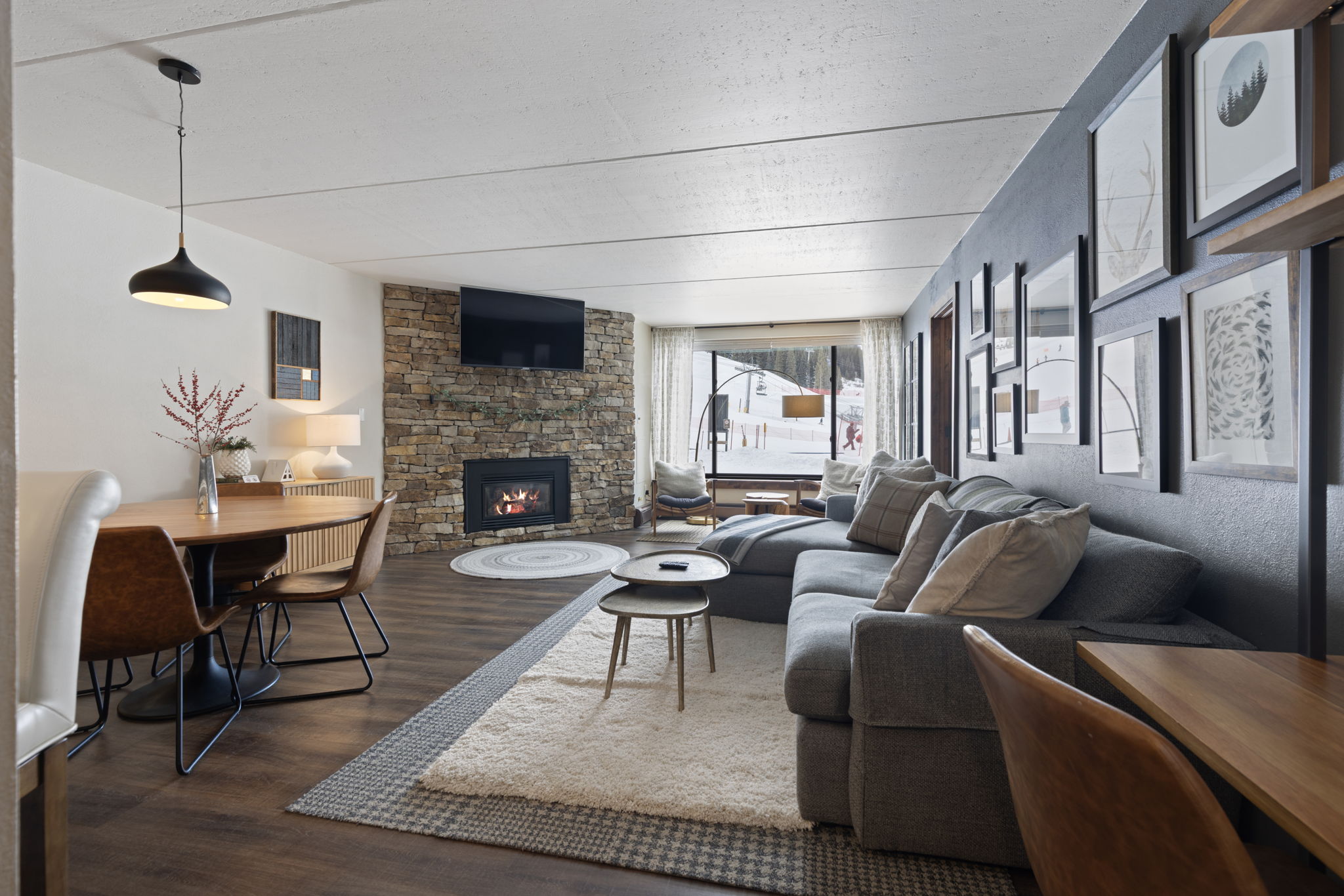This open-concept living and dining area features a cozy, mountain-inspired aesthetic with a prominent stone fireplace and a large sectional sofa. The space is anchored by a plush area rug and illuminated by a modern pendant light over the dining table, while a large window provides a scenic view of a snowy ski slope. The warm wood flooring and dark accent wall with gallery art create a sophisticated yet inviting atmosphere perfect for a mountain retreat.