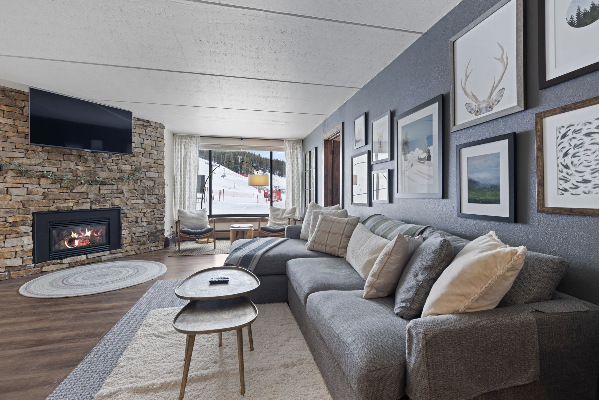 This cozy, mountain-inspired living room features a striking floor-to-ceiling stone fireplace with an integrated television and a large sectional sofa. The space is anchored by a plush area rug and layered coffee tables, while a large window provides a picturesque view of a snowy ski slope. The dark accent wall adorned with a gallery of nature-themed artwork adds a sophisticated, rustic charm to the inviting interior.