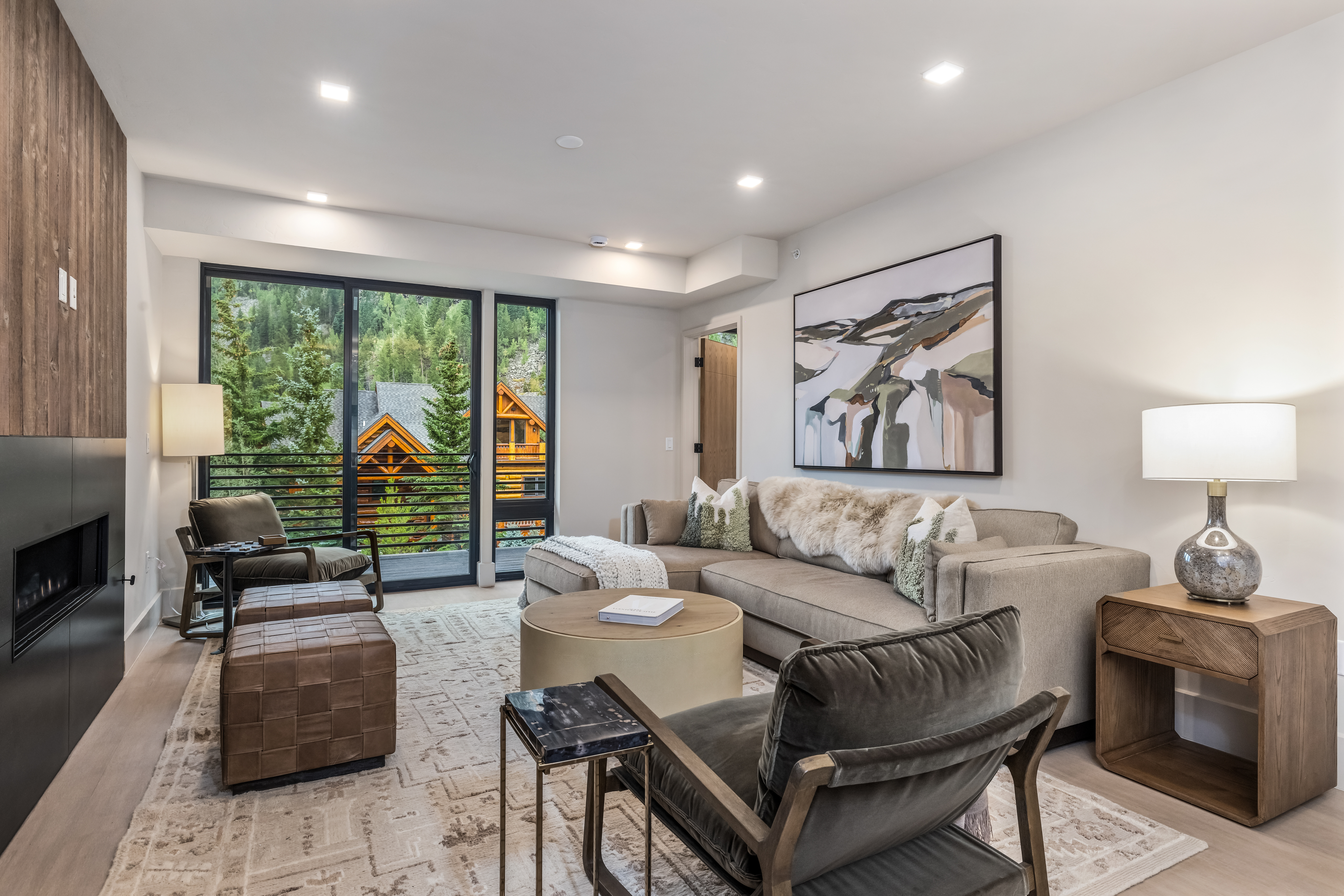 This is a well-lit living room featuring a modern design with a neutral color palette. The room includes a large sectional sofa, a round ottoman, and comfortable armchairs, creating a cozy and inviting atmosphere. A large window offers a view of the surrounding trees and a glimpse of a nearby house, enhancing the connection to the outdoors.
