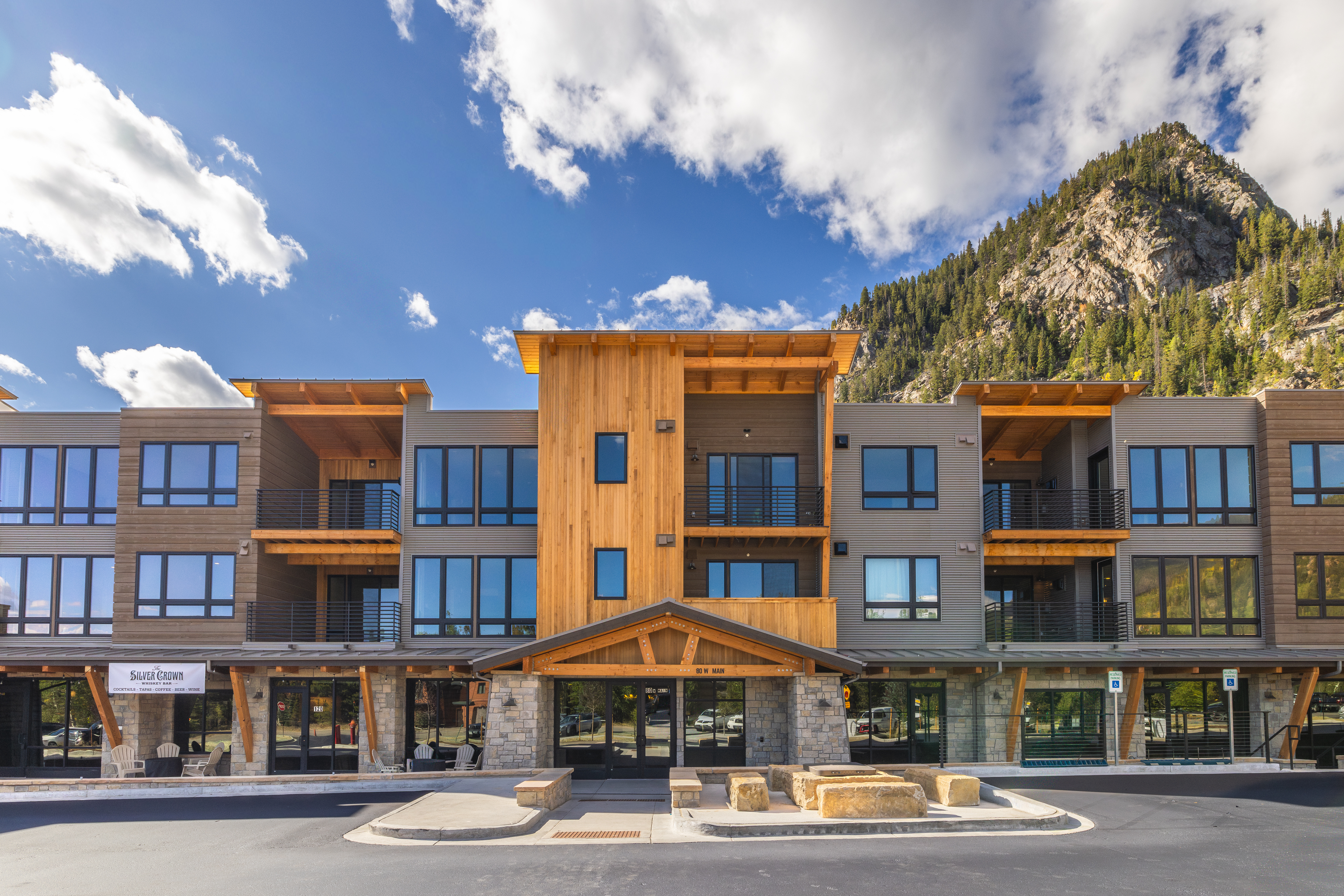 This is a front exterior view of a modern multi-story building, possibly a condominium or apartment complex. The building features a mix of wood and metal siding, large windows, and balconies. The entrance is accented with stone and wood details, creating a welcoming and upscale impression, with a mountain in the background.