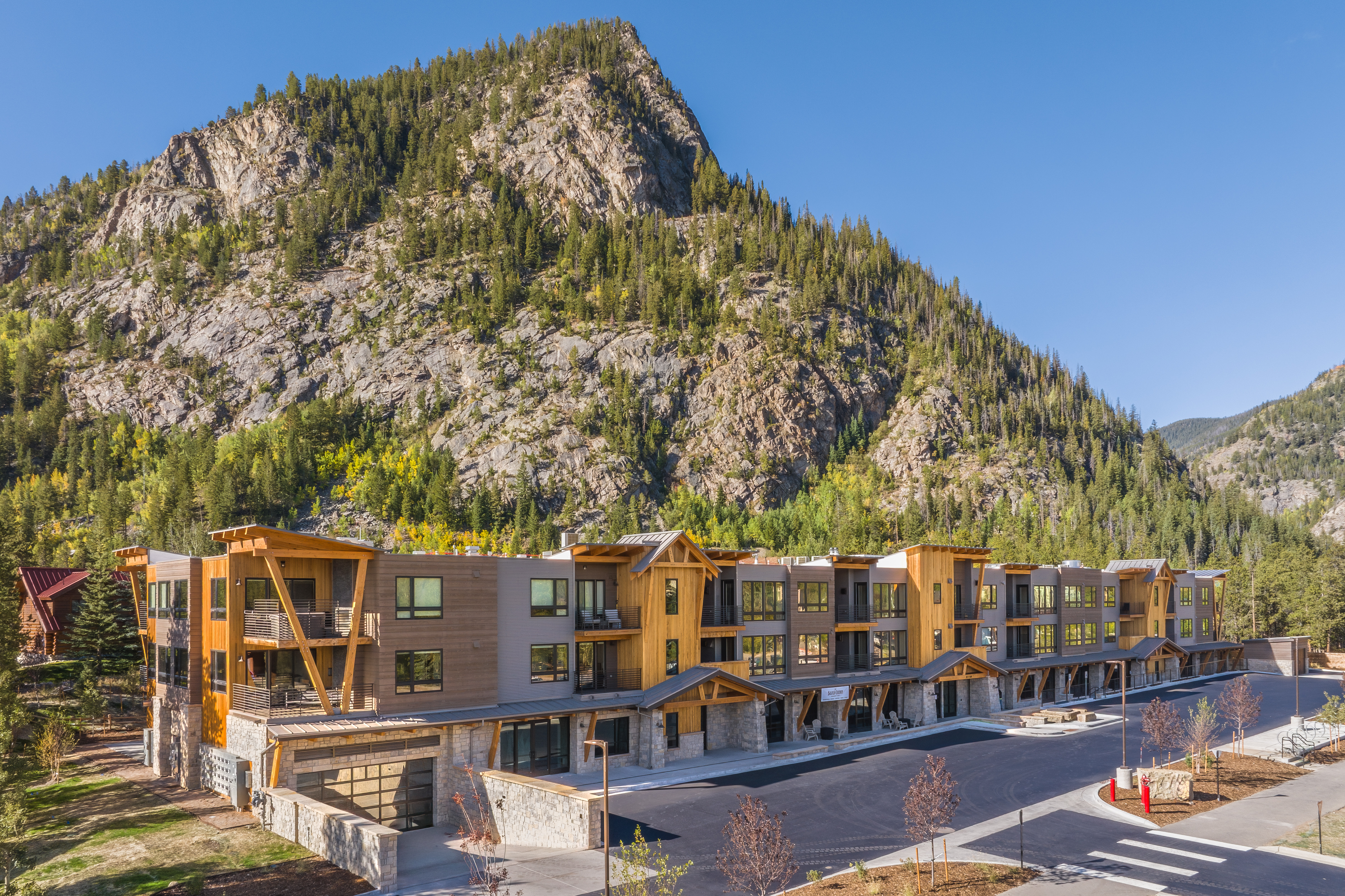 This is a front exterior view of a modern multi-family residential building nestled against a mountain backdrop. The building features a mix of wood and gray siding, with balconies and large windows. The landscaping is well-maintained, and a paved road leads to the entrance, creating a welcoming and upscale impression.