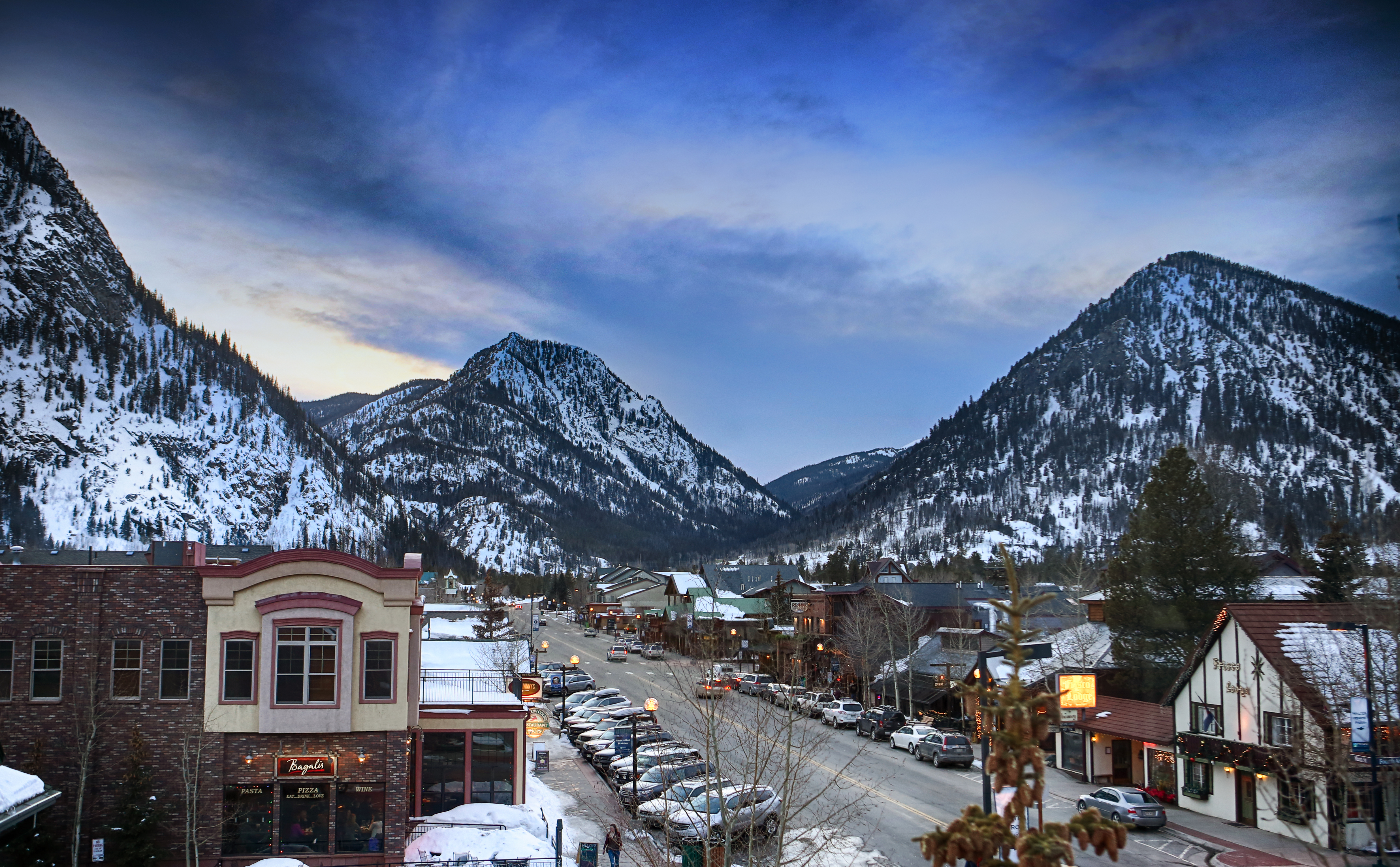 This aerial view showcases a charming mountain town nestled between snow-capped peaks under a twilight sky. The main street is lined with quaint buildings, many with architectural details, and cars parked along the sides, suggesting a vibrant community. The scene evokes a sense of serenity and the allure of mountain living.