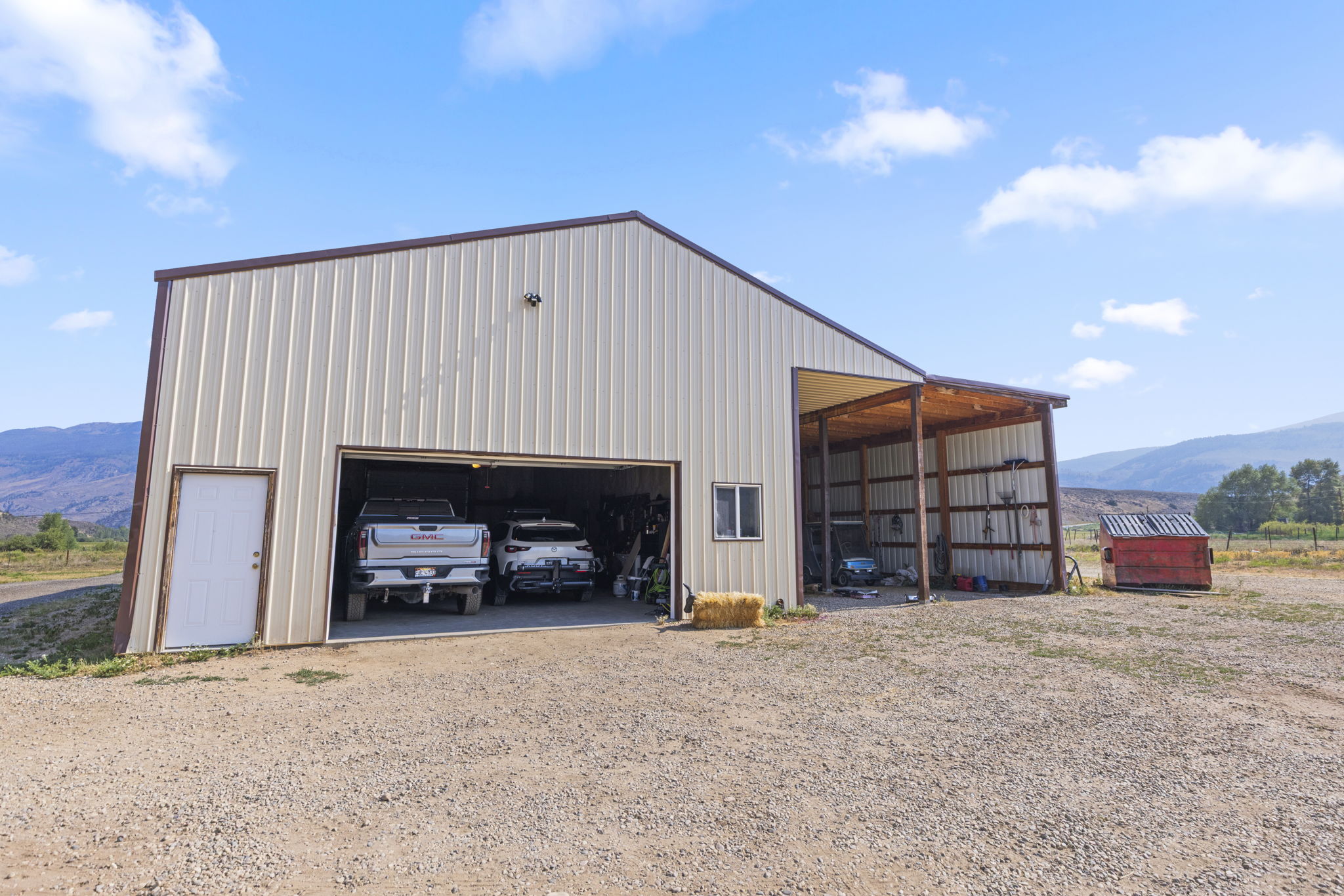 This image features a large, metal-sided utility building or workshop set against a clear blue sky and mountain backdrop. The structure includes a wide, open garage bay housing two vehicles, a separate pedestrian door, and an attached open-air carport area on the right. A gravel driveway leads up to the building, which is situated in a rural, open landscape.