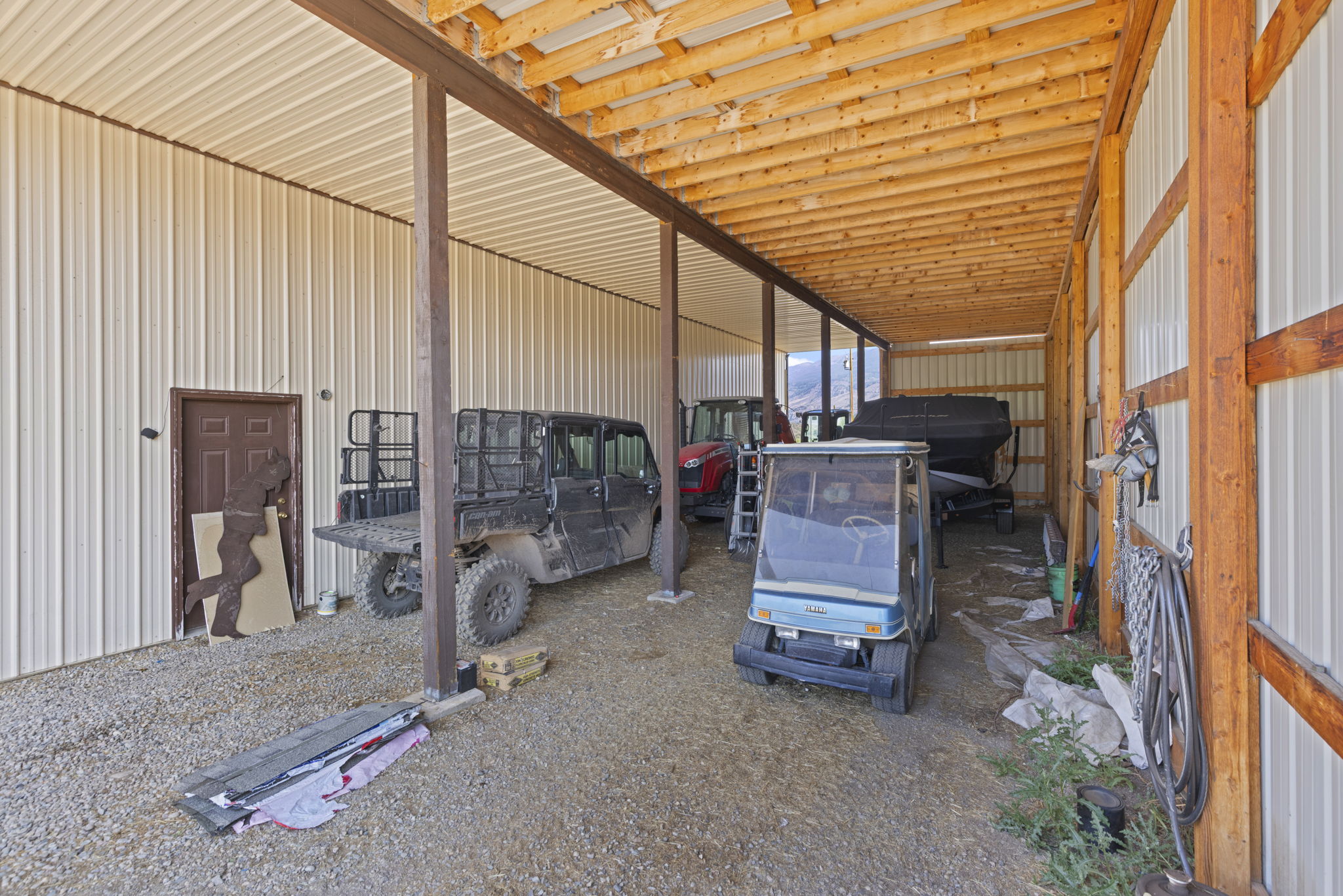 This image shows a large, open-sided metal storage structure or pole barn designed for housing vehicles and equipment. The space features a gravel floor, exposed wooden rafters, and metal siding, providing a functional and utilitarian storage area. Parked inside are a utility vehicle, a tractor, and a golf cart, with a boat trailer visible in the background, highlighting the structure's capacity for versatile storage.