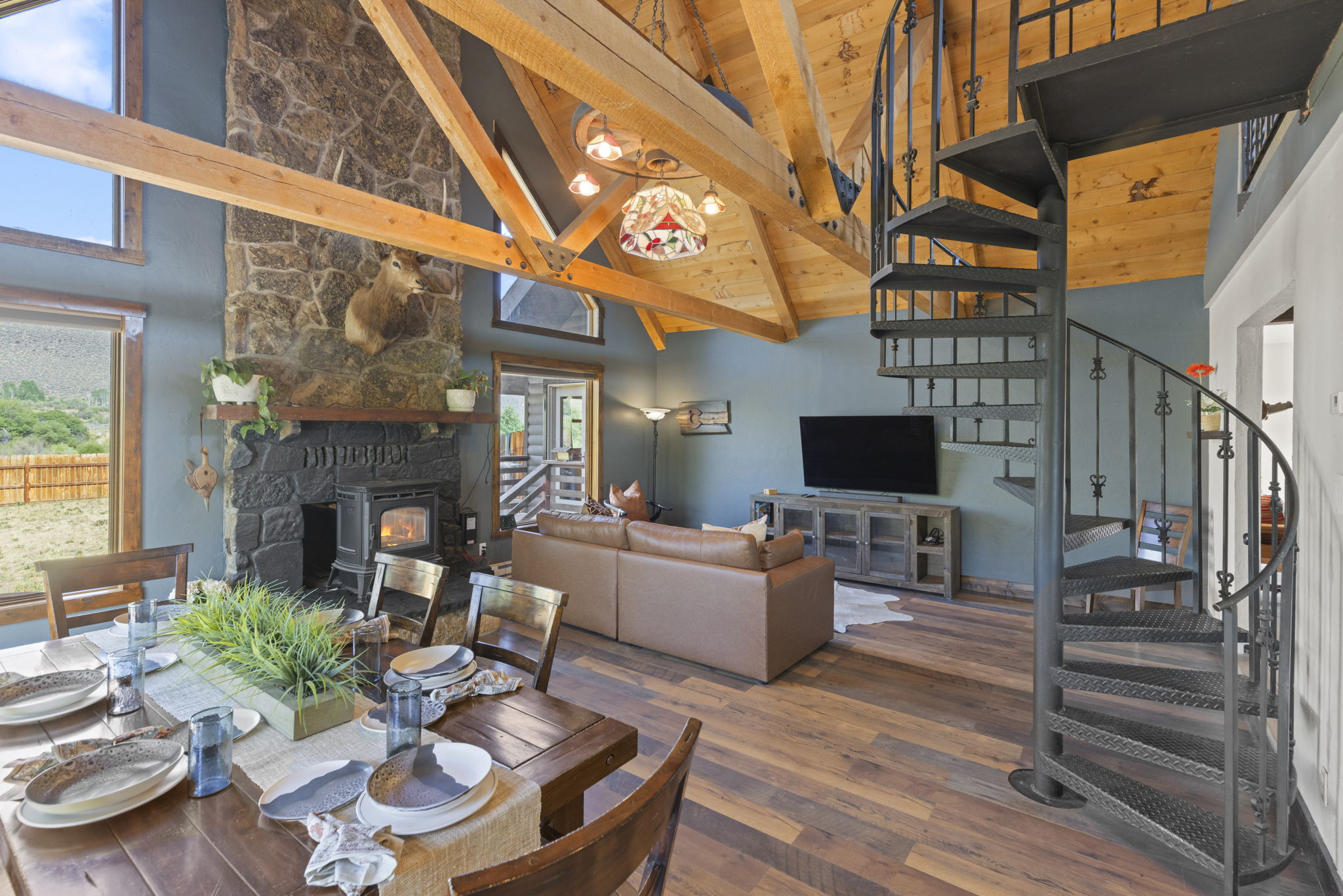This open-concept living and dining area features a rustic aesthetic with exposed wooden beams and a vaulted ceiling. A prominent stone-clad chimney breast serves as a focal point, complemented by a leather sofa, a wooden dining table, and a spiral staircase leading to an upper level. Large windows allow natural light to fill the space, highlighting the warm wood flooring and the cozy, cabin-inspired atmosphere.
