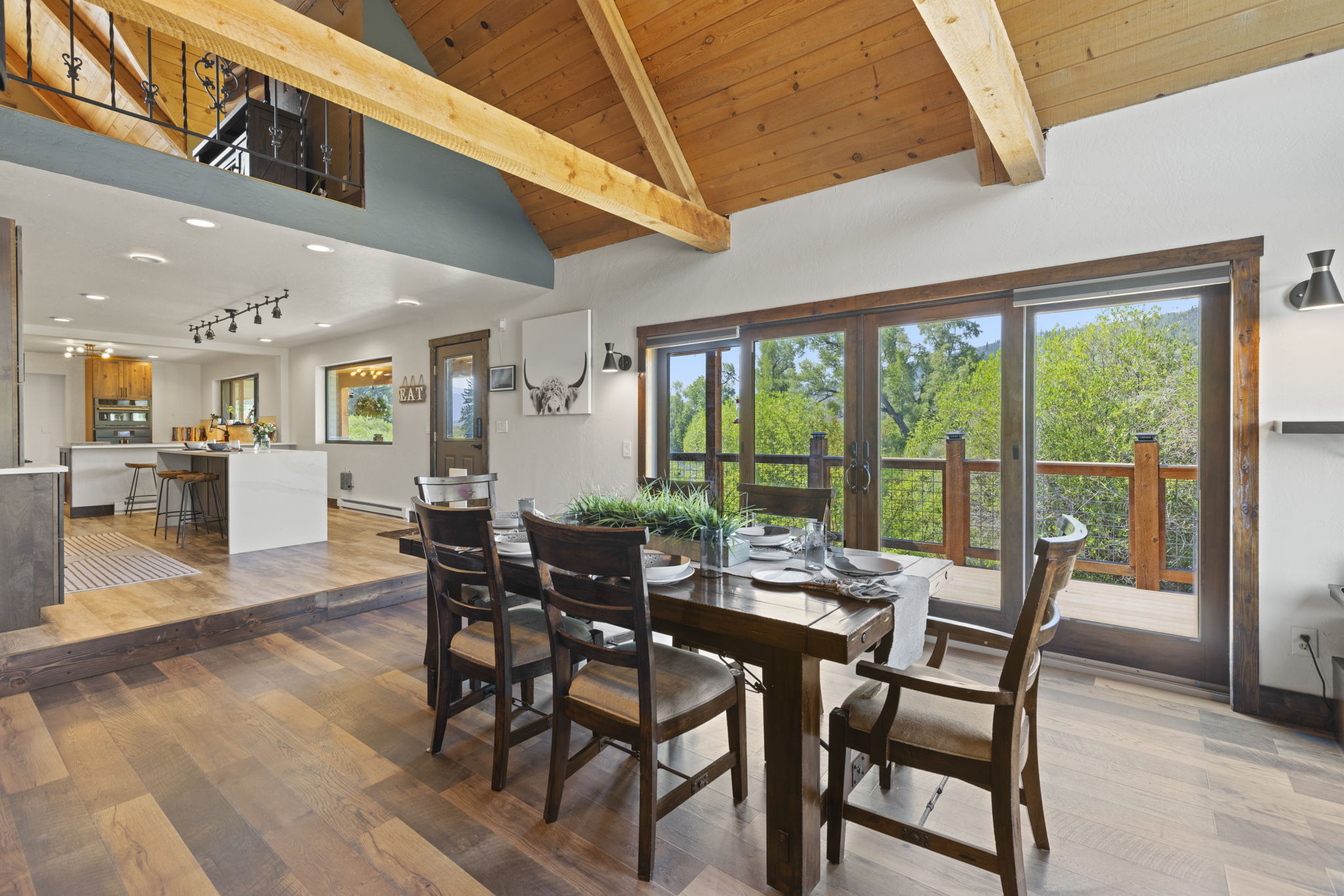 This open-concept dining area features a rustic wooden table surrounded by matching chairs, set beneath a dramatic vaulted ceiling with exposed timber beams. Large sliding glass doors provide a seamless transition to an outdoor deck, filling the space with natural light and scenic views. The room flows directly into a modern kitchen, creating an airy and cohesive living space with warm wood flooring throughout.