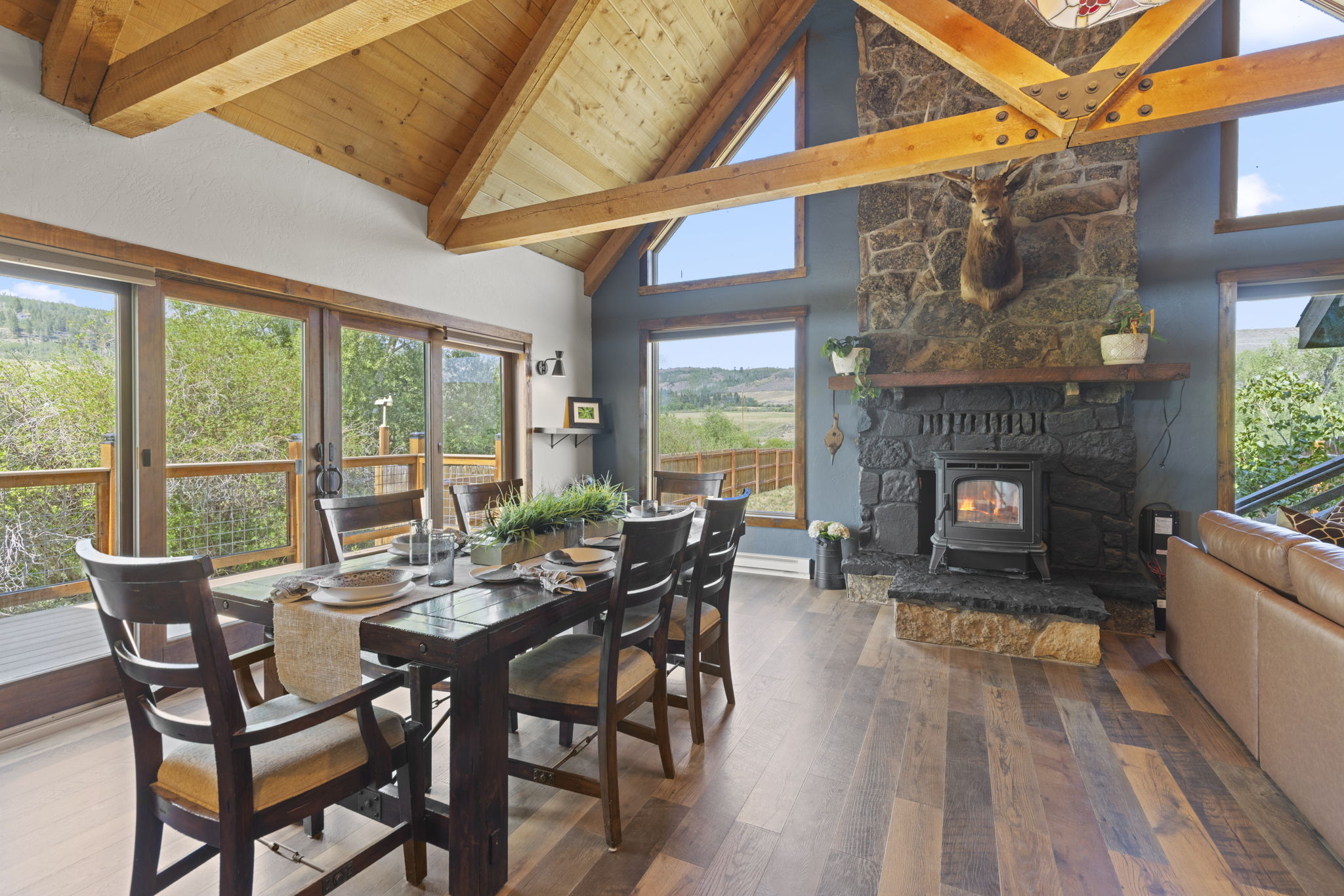 This rustic dining area features a large dark wood table surrounded by matching chairs, set against a backdrop of a stone-clad chimney breast with a built-in wood stove. The room is defined by its vaulted wood-paneled ceiling, exposed timber beams, and expansive windows that provide abundant natural light and scenic views of the surrounding landscape. The combination of warm wood tones, dark stone, and blue-toned walls creates a cozy, mountain-lodge atmosphere.