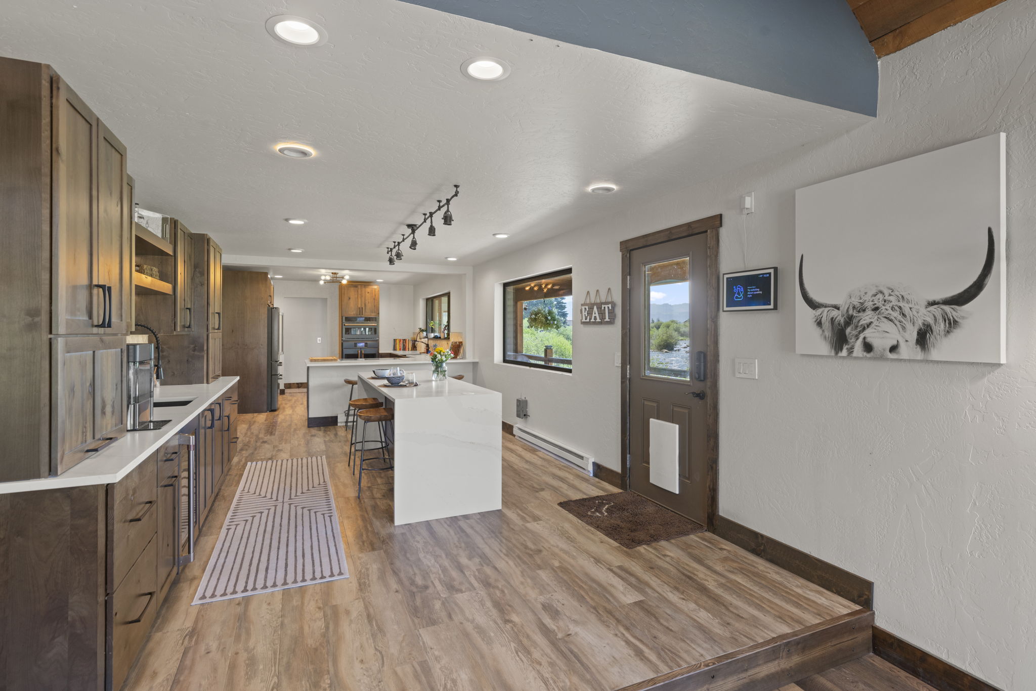 This modern kitchen features warm-toned wood cabinetry paired with light-colored countertops and a central island with bar seating. The space is illuminated by recessed lighting and a track light fixture, while a large window provides a view of the outdoors. A decorative 'EAT' sign and a large wall print of a highland cow add character to the open-concept layout, which transitions into a slightly elevated entryway.