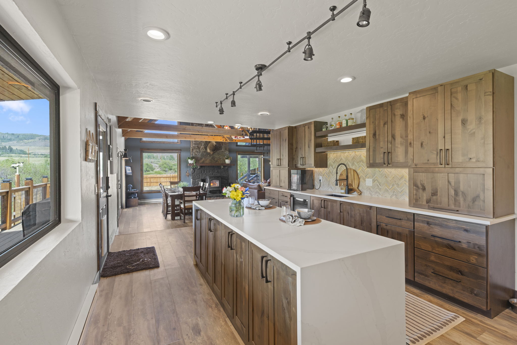 This open-concept kitchen features warm-toned wood cabinetry, a large central island with a light-colored countertop, and a herringbone-patterned backsplash. The space flows seamlessly into a dining area with a stone-clad chimney breast and large windows that provide a view of the surrounding landscape. The perspective is from the kitchen looking toward the dining area, highlighting the rustic, inviting atmosphere of the home.