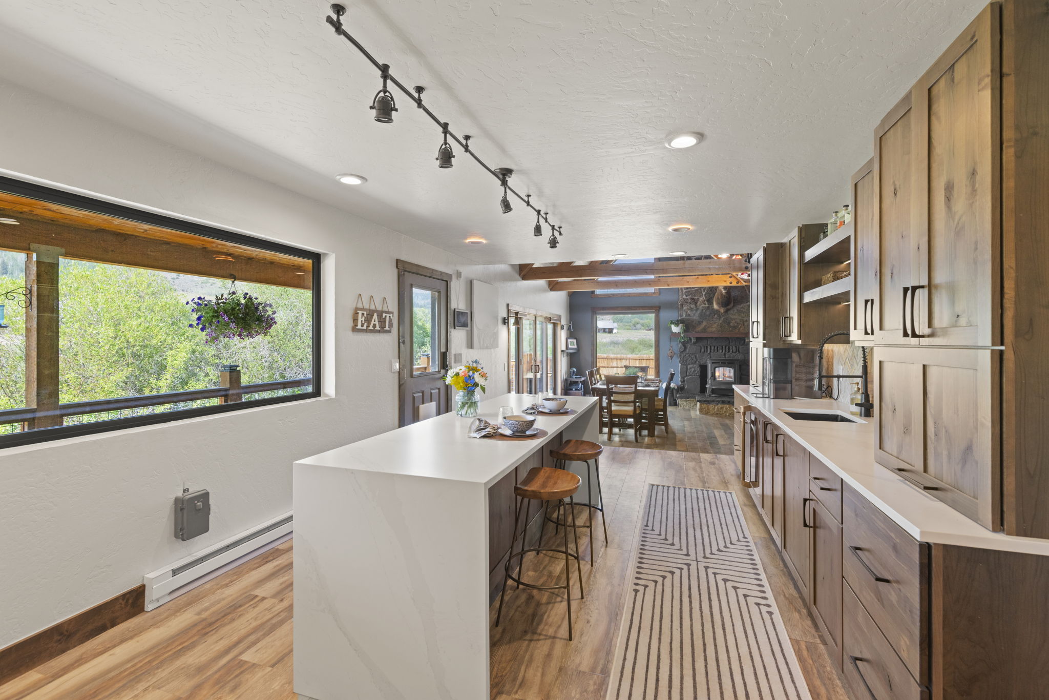 This bright, open-concept kitchen features a large central island with a light-toned stone countertop and two wooden stools. The space is anchored by warm-toned wood cabinetry and light wood-look flooring, with a long track lighting fixture overhead. A large window provides a scenic view of the outdoors, while the perspective looks through to a dining area and a stone-clad fireplace in the adjacent room.