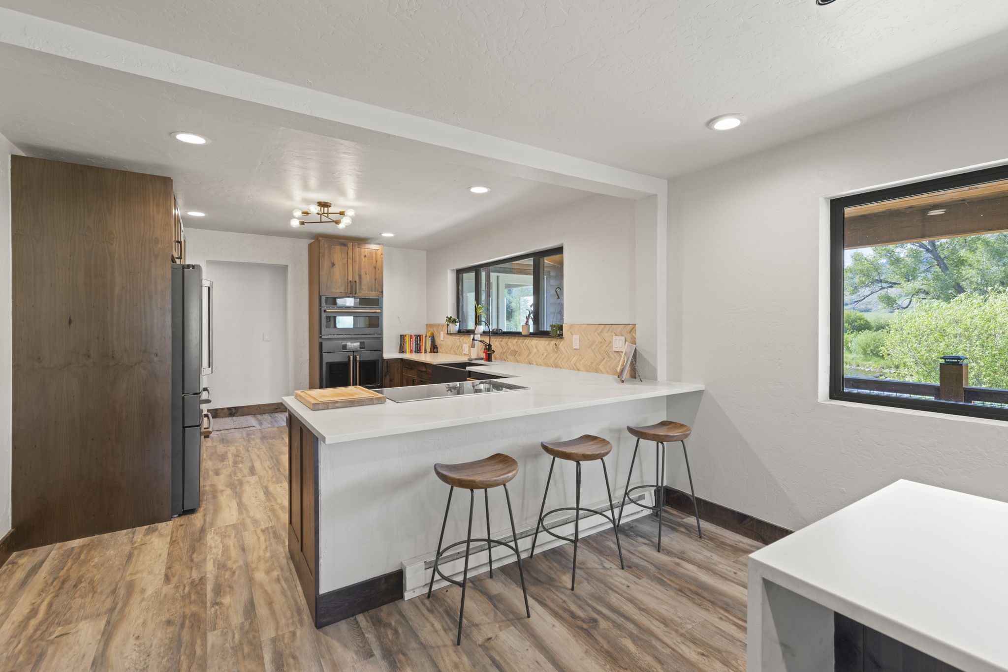 This modern kitchen features a spacious island with a white countertop and three wooden bar stools, creating a functional breakfast bar area. The space is accented by warm wood-toned cabinetry, a herringbone-patterned backsplash, and a large window that provides a scenic view of the outdoors. The open-concept layout flows seamlessly into the adjacent living area, highlighted by light-colored flooring and recessed lighting.