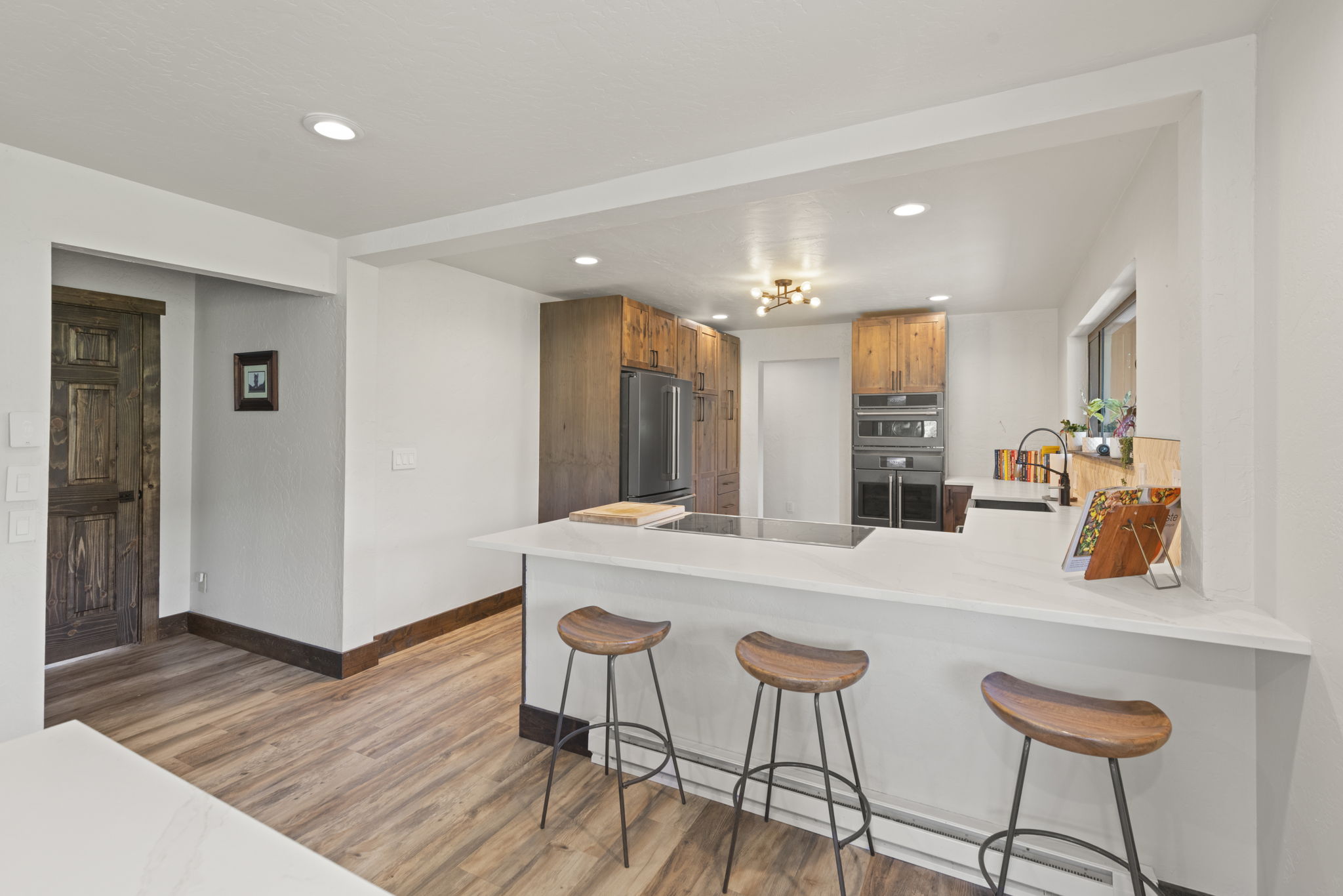 This bright, open-concept kitchen features a large peninsula with a white countertop and three wooden bar stools, perfect for casual dining. The space is anchored by warm-toned wood cabinetry, a built-in oven, and a sleek cooktop, creating a balanced blend of rustic and modern aesthetics. The perspective captures the flow from the kitchen into the adjacent hallway, highlighting the wood-look flooring and clean, neutral wall palette.