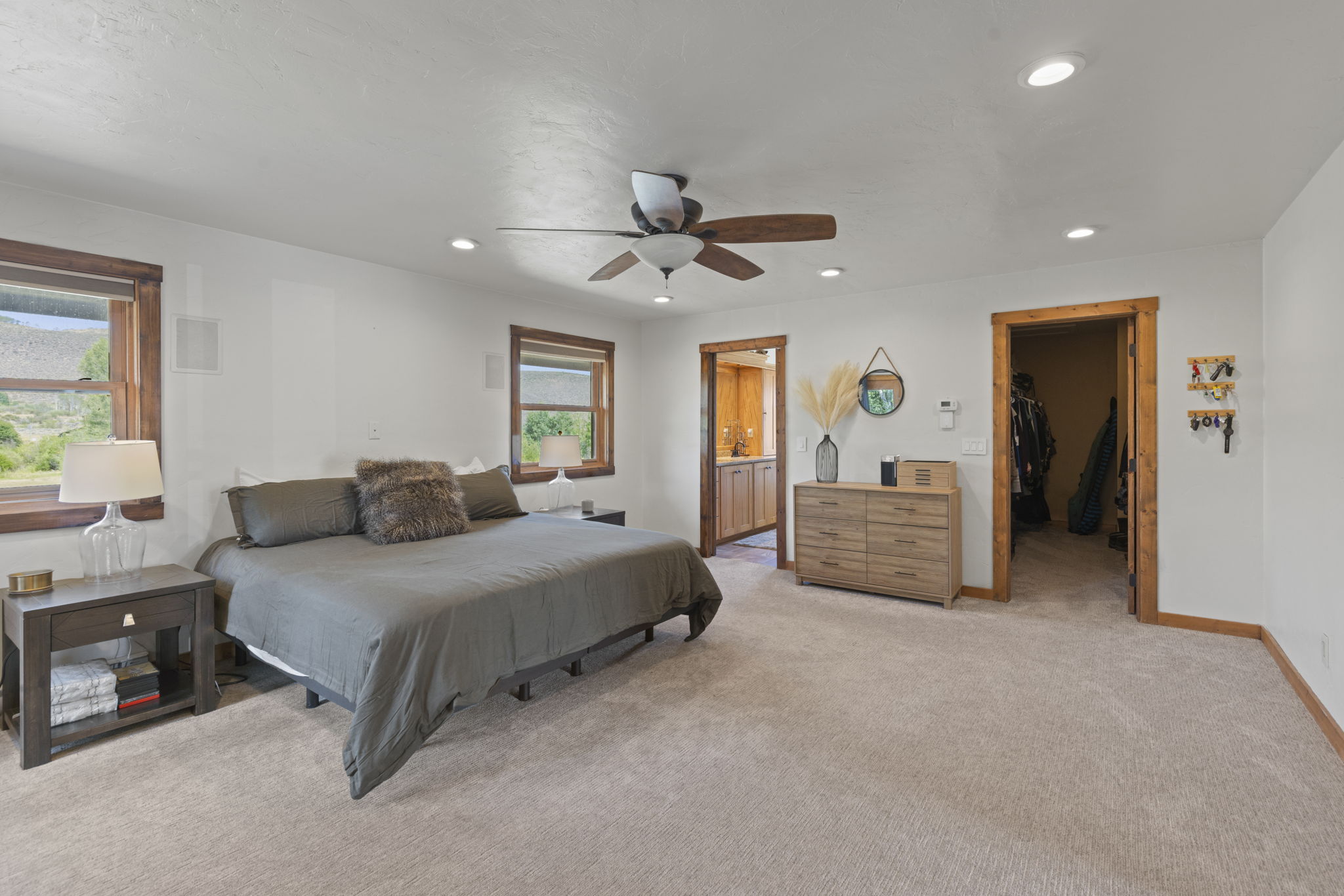 This spacious primary bedroom features neutral walls, plush carpeting, and a large bed centered against the wall with two matching nightstands. The room includes a ceiling fan, recessed lighting, and two windows that provide natural light and views of the outdoors. A wooden dresser sits between the doorways leading to an en-suite bathroom and a walk-in closet, creating a functional and well-organized layout.