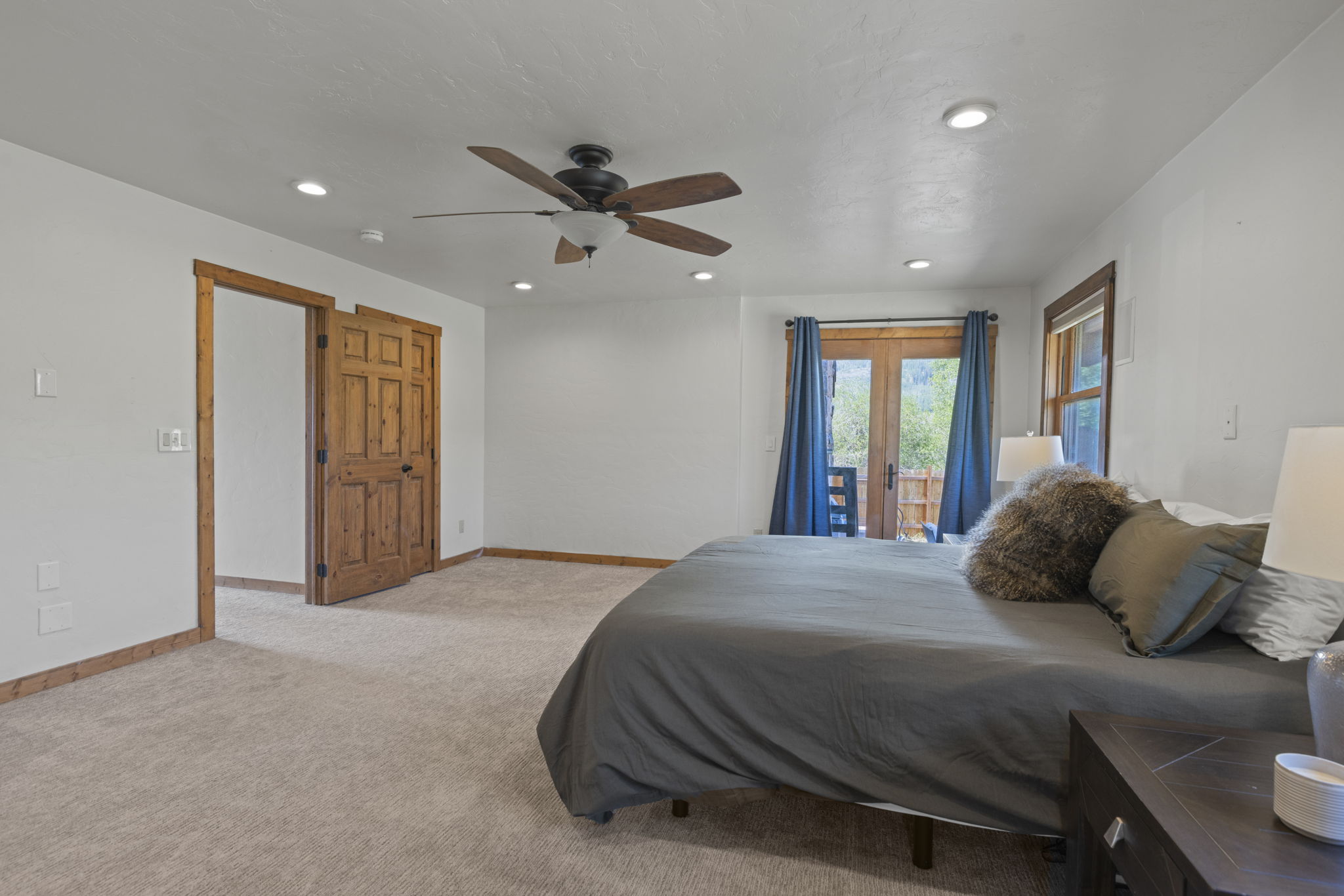 This spacious primary bedroom features neutral carpeting, white walls, and a ceiling fan with a light fixture. A large bed with a dark gray comforter and a textured accent pillow is positioned on the right, while a set of wooden double doors leads to an adjacent room. French doors with blue curtains provide access to an outdoor space, allowing natural light to fill the room.